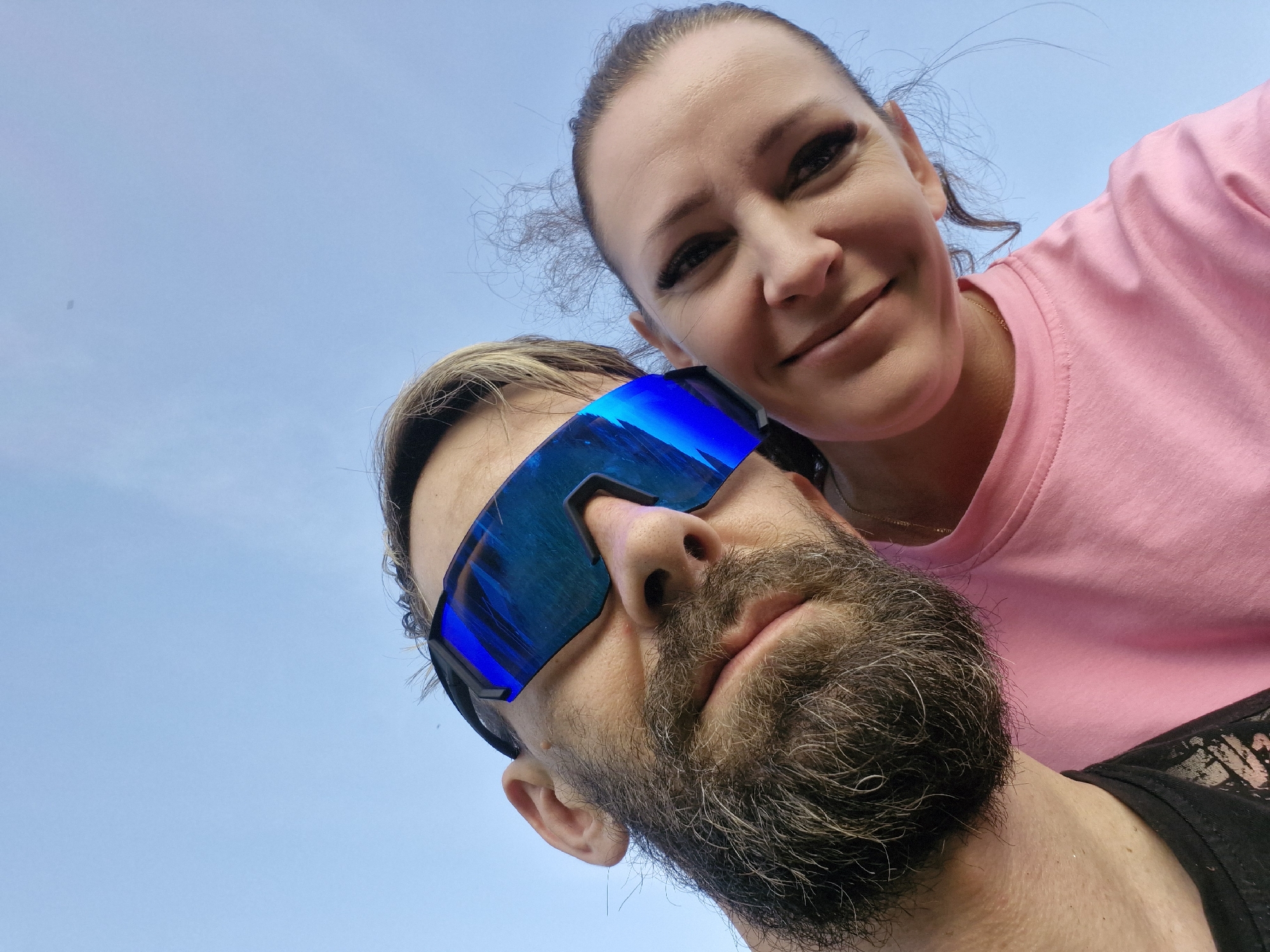 An outdoor, low-angle, close-up selfie shows a man and a woman against a clear blue sky. The man is on the left, facing the camera and wearing large, blue mirrored sunglasses that cover most of his face. He has a beard and what appears to be light brown hair. He is wearing a black t-shirt with some white detailing. The woman is on the right, looking at the camera with a smile. She has dark hair pulled back and is wearing a pink t-shirt. Her makeup includes dark eyeliner and mascara. The image was taken outdoors with the sky as the backdrop, suggesting it could be in an open area like a park or a street in Марковский, Russia.