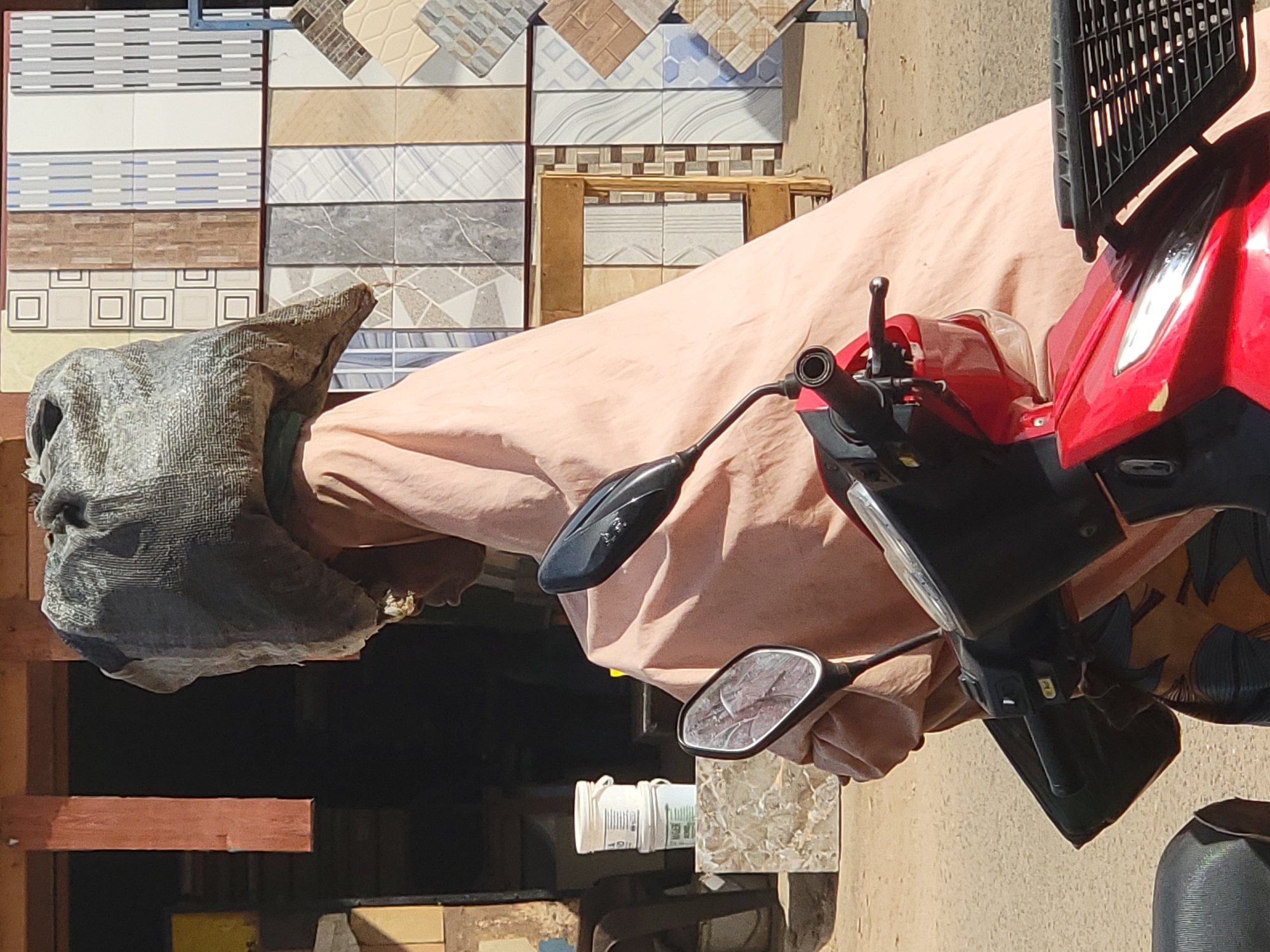 A person is sitting on a red motorcycle parked on a street in Maiburiji, Nigeria. The motorcycle is draped with a light peach-colored cloth, and the rider is wearing a gray head covering. In the background, there are stacks of patterned tiles. A bottle of liquid is nearby. The scene appears to be outdoors during the daytime.