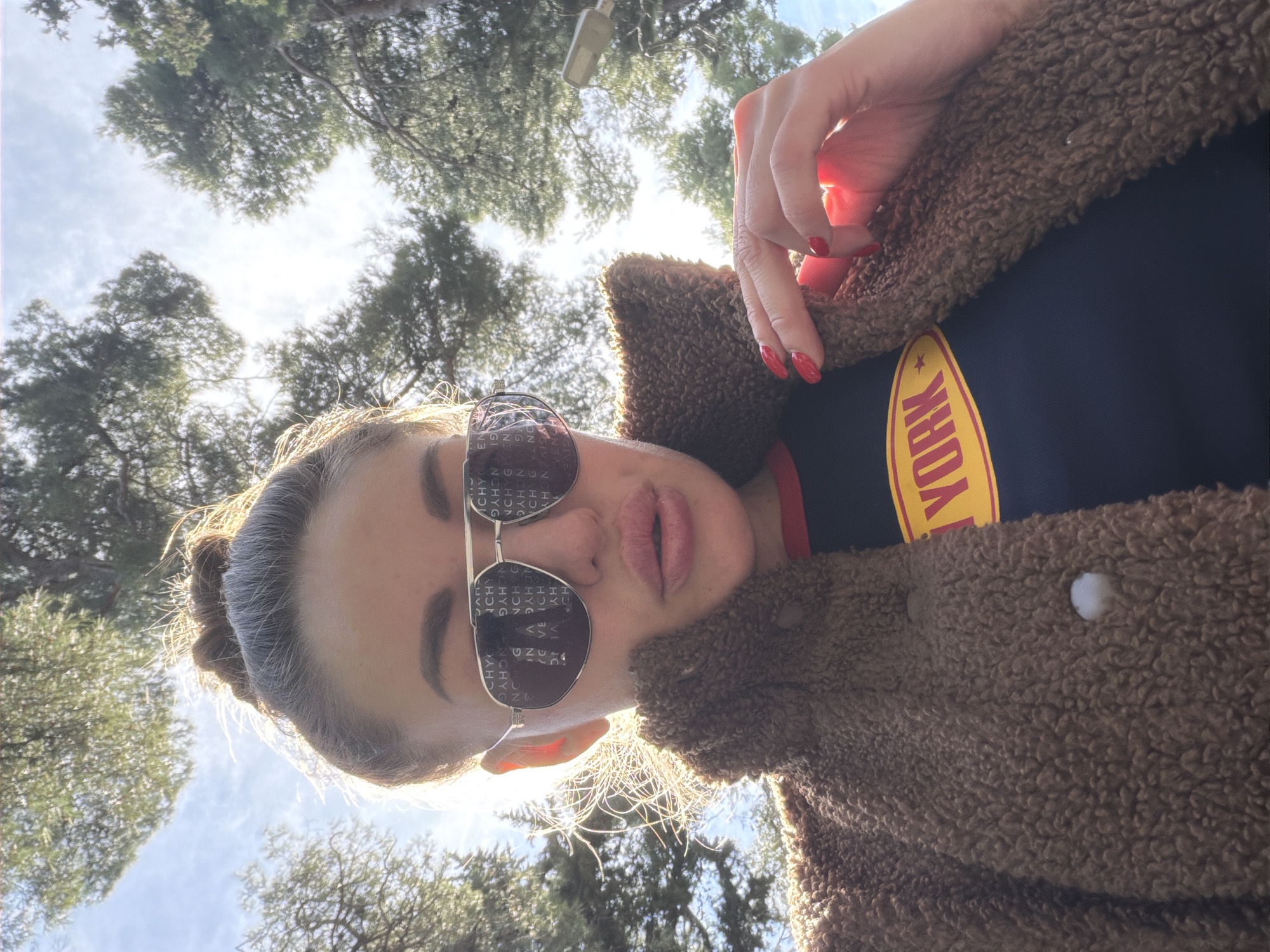 A close-up, low-angle selfie captures a person looking upwards outdoors in Bar, Montenegro, during bright daylight. The background features a partially cloudy sky and numerous green trees, suggesting a park or natural setting.
The subject, likely female, is wearing large aviator-style sunglasses. The lenses of the sunglasses prominently display repeated text, which includes "GIVENCHY," "NCHYG," "CHYC," "VEIC," and "ENCHYG." She is dressed in a textured, brown teddy or sherpa-style jacket. Beneath the jacket, a dark top is visible, featuring a yellow design with a star and the word "YORK," presumably part of "NEW YORK."
Her hand, adorned with bright red nail polish, is visible near her shoulder, subtly adjusting her jacket. Her hair is pulled back, with sunlight illuminating loose strands around her face and scalp. The overall scene is bright and well-lit by natural sun, creating some lens flare effects. The person's expression is neutral, with her lips slightly parted.