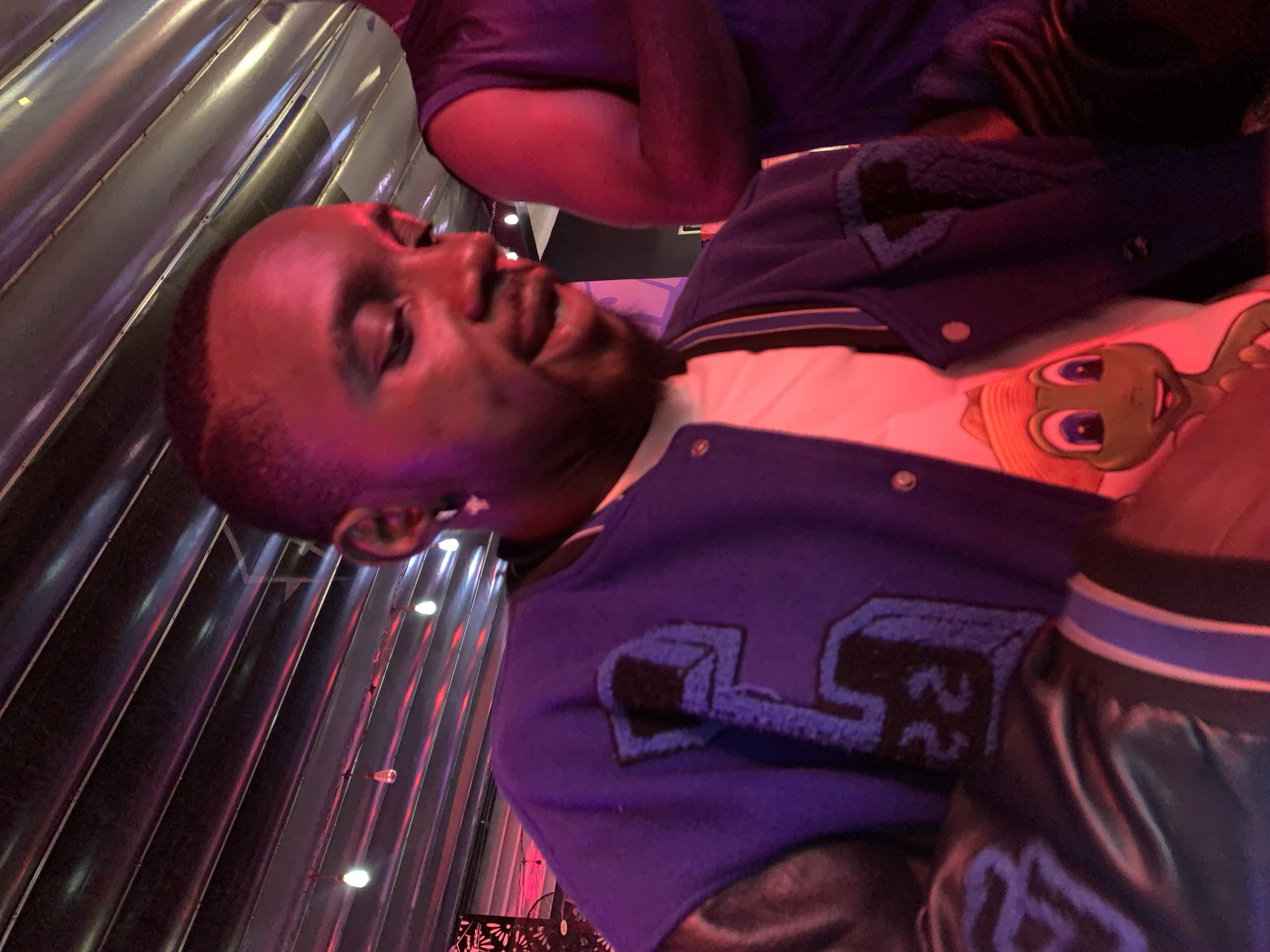 A dark-skinned man wearing a purple and blue letterman jacket with "5" and "22" on it, and a white t-shirt with a cartoon character on it, is looking down and to the right. He has a gold earring in his left ear. The background is a dark ceiling with exposed metal beams and purple and pink lighting. Another person in a dark purple shirt is partially visible to the right. The man appears to be in a dimly lit, possibly indoor venue in the city of Iwo, Osun State, Nigeria.