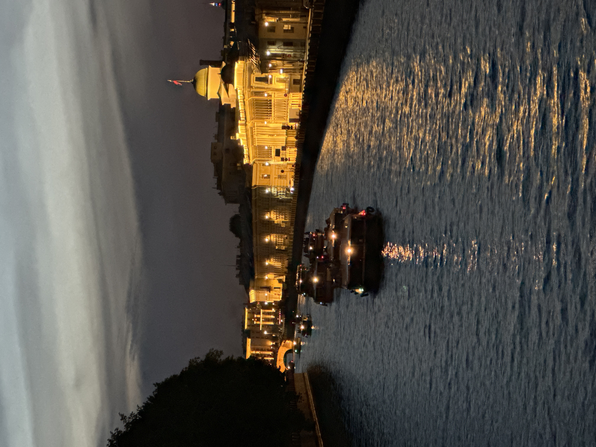 This image depicts a nighttime scene along a canal in Saint Petersburg, Russia, likely the Moika River, given the architectural style and specific landmark. The sky overhead is dark grey with prominent horizontal cloud formations, indicating late twilight or early night.
In the foreground, the dark, rippled surface of the canal occupies the lower half of the frame. Streaks of orange and yellow light are reflected on the water, originating from the illuminated buildings and boats. A procession of approximately seven tour boats, each equipped with internal and external lighting, navigates the canal from the foreground towards the background. Passengers are visible on the decks of these boats.
The left bank of the canal is partially obscured by a dark, silhouetted mass of trees in the immediate foreground. Further along the left bank, an illuminated stone arch bridge with a warm yellow glow spans the canal in the middle ground, connecting to a distant line of illuminated buildings.
The right bank of the canal features a continuous row of brightly illuminated historical buildings. The most prominent structure, located on the right side of the frame, is a large, ornate building with neoclassical elements, distinguished by a golden dome topped with a flagpole from which a red, white, and blue striped flag (the Russian tricolor) flies. This building is identifiable as the former Senate and Synod building, now housing the Constitutional Court of Russia. The buildings along this bank are uniformly lit with warm, yellow-orange light, highlighting their architectural details. The overall environment is urban, characterized by artificial light against the dark sky and water.