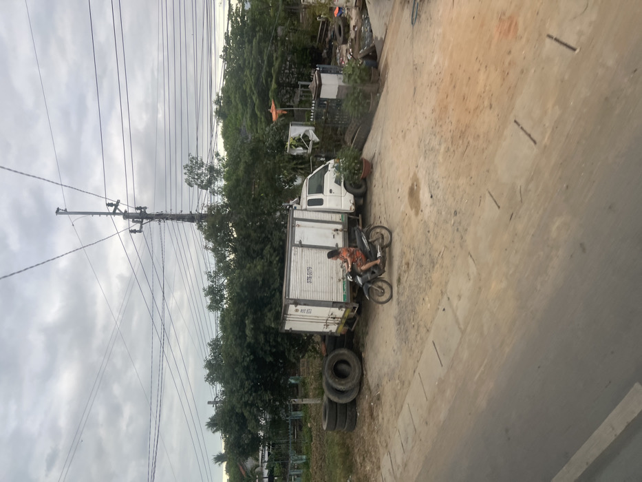 An outdoor, daytime scene in Ninh Tây, Vietnam, captured under an overcast sky. The foreground features a paved road with a concrete curb separating it from an expansive, uneven dirt and gravel area. In the midground, a woman in an orange patterned dress is seated on a dark gray scooter, oriented right, with her right foot resting on the ground, indicating a stationary or slow-moving posture. To the left of the scooter, a stack of five large, dark vehicle tires is present. Behind the woman and the tire stack, a white box truck, marked with "57S-3179" on its side, is parked. The truck's front cab is white. To the right of the truck, an unpaved incline leads upward past a low dark fence and a decorative pillar, revealing additional tires, discarded materials, and potted plants. The background is dominated by dense, lush green foliage and trees, partially obscuring what appear to be residential structures on the far left. A tall utility pole with multiple intersecting overhead power lines extends vertically into the gray, cloud-covered sky.