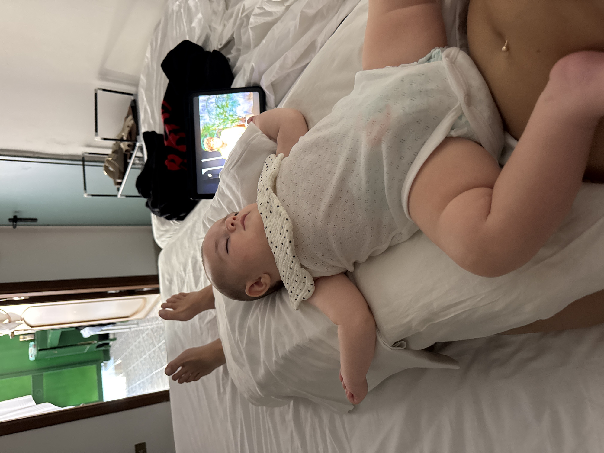 The media file shows a close-up, overhead view of a baby lying on a white pillow on a bed. The baby is wearing a white onesie with a dotted bib. The baby's eyes are closed, and their face is relaxed, suggesting they are asleep or resting. The baby's legs and arms are visible, and one of the baby's feet is positioned near the viewer. To the right of the baby, an adult's torso is visible, with a navel piercing. The adult's skin tone suggests they are likely of Mediterranean descent.
In the background, a tablet device is propped up, displaying an image with animated characters and a nature scene. There are also some dark clothing items on the bed. Further in the background, a rack with items hanging on it is visible, as well as a glimpse of a room with a green shelf unit. The overall setting appears to be indoors, possibly a bedroom, with natural light entering from a window or doorway.
The image is oriented sideways. The presence of the navel piercing and the skin tone hint at a warmer climate. Given the location context of Isola delle Femmine, Italy, the time of day is likely during daylight hours. The overall mood of the image is peaceful and intimate, focusing on the rest of the baby. There is no visible text in the image.