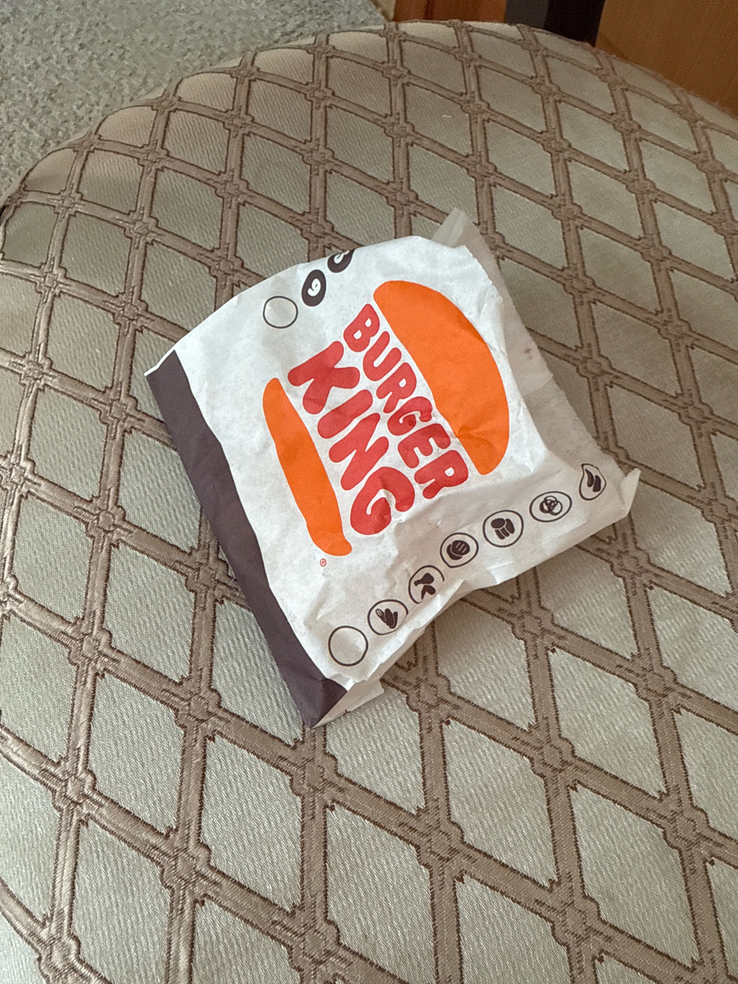 A crumpled Burger King paper bag rests on a textured, light-colored ottoman with a diamond-shaped pattern. The bag is white with the orange and red Burger King logo visible. Small black icons are printed along the bottom edge of the bag. The background is a muted, textured surface. The image is taken from a slightly elevated angle, looking down at the bag. There are no people or animals in the image.