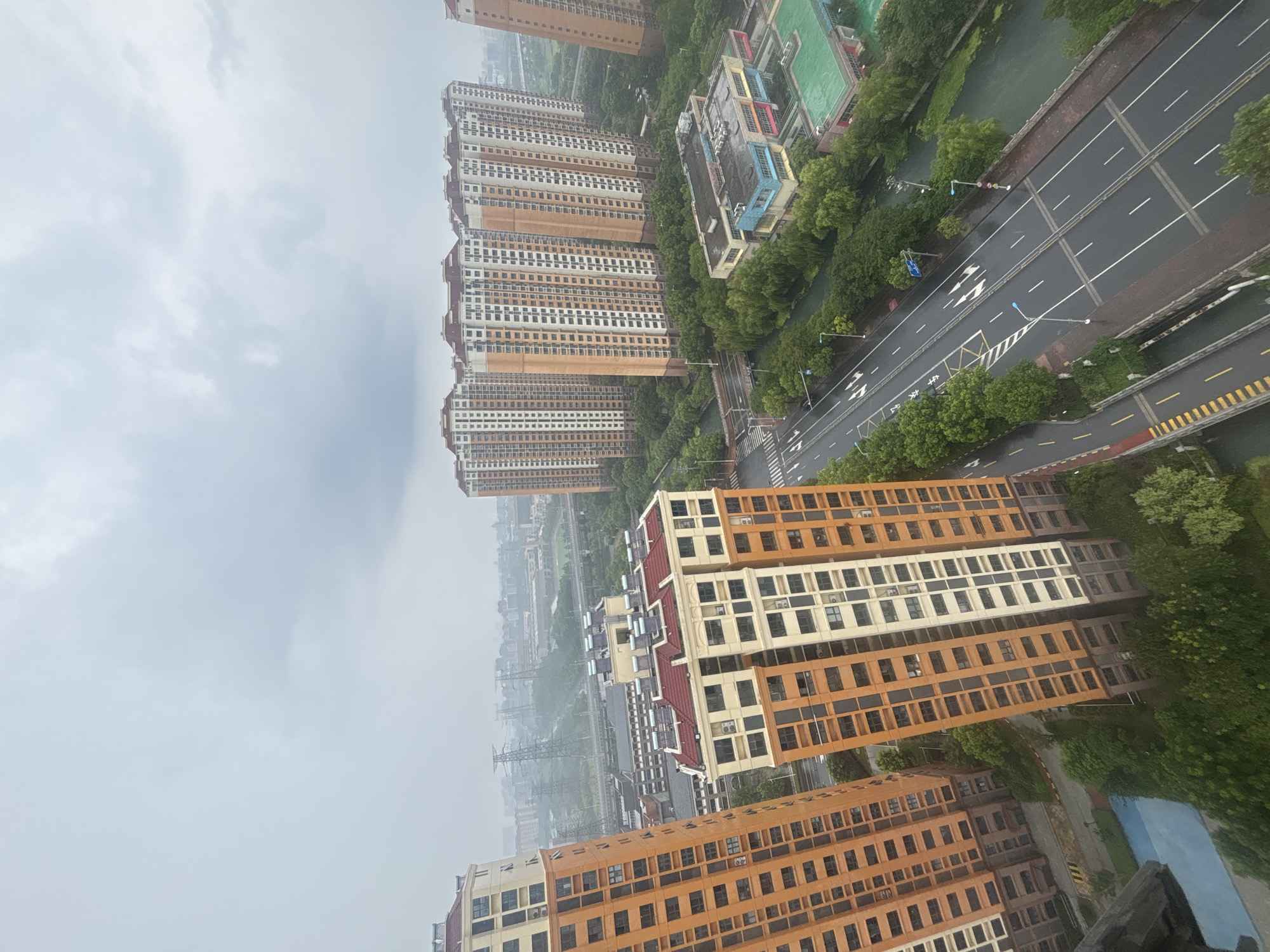 This is a high-angle, medium shot of an urban environment on a cloudy day. The scene appears to be the Suzhou Industrial Park in China, based on the architecture and infrastructure. In the foreground, two tall, multi-story residential buildings are visible, predominantly orange and cream colored, with numerous windows. One building is closer to the viewer on the left, and another is slightly further away to the right, with more buildings of similar design stretching into the background. A wide, wet road with multiple lanes and directional markings runs through the middle ground, with streetlights and some trees lining its edges. On the right side of the road, there are some smaller, low-rise buildings, including what appears to be a school with colorful sections and a green sports field. A body of water, possibly a canal or river, runs alongside the road and between some of the buildings, lined with lush green trees. In the background, more buildings and industrial structures, including tall power lines and towers, are visible through a hazy atmosphere. The sky is overcast with heavy gray clouds. No people are discernible in the image. The overall impression is of a dense, modern urban development, possibly after or during rainfall.