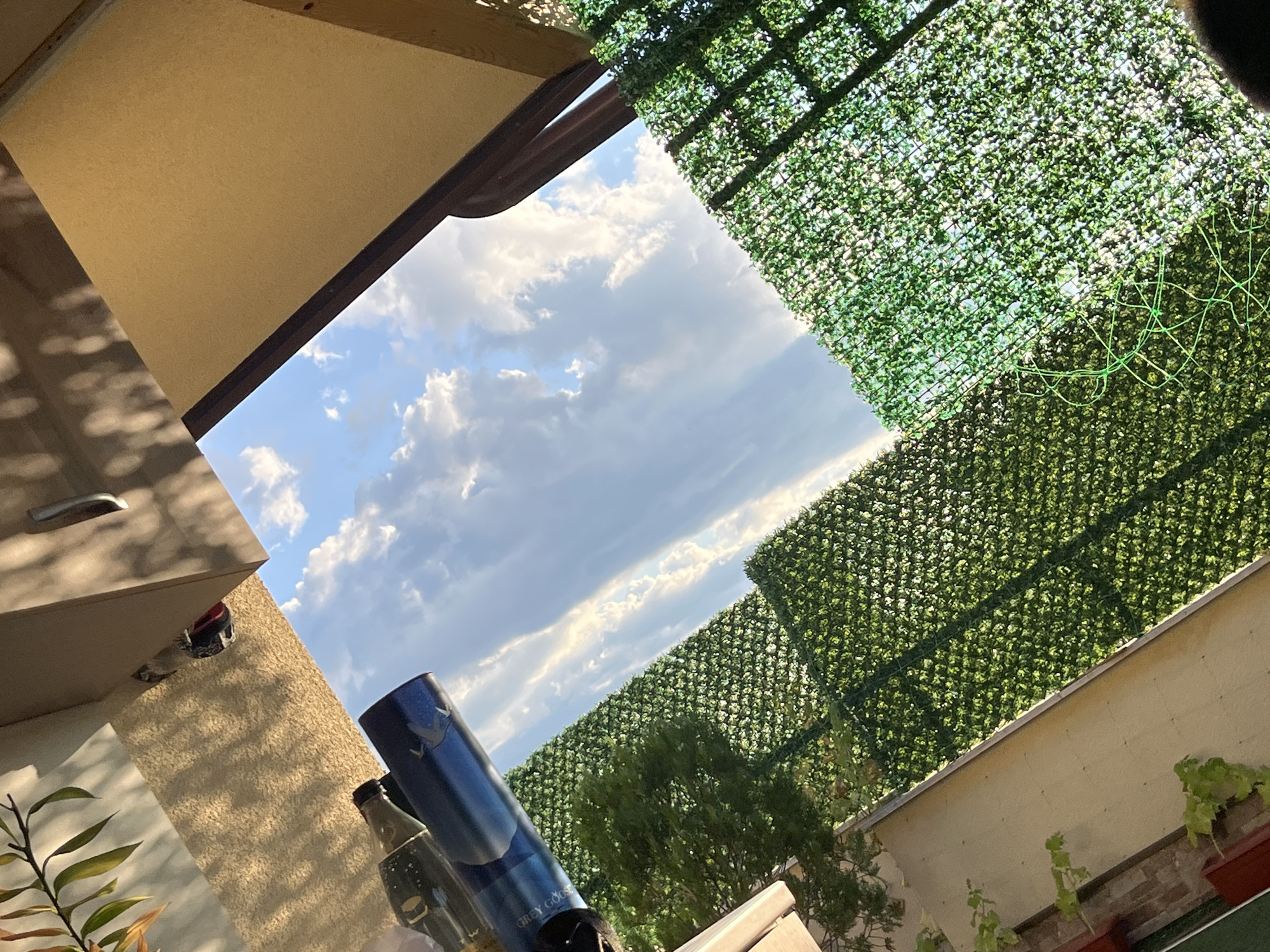 The image shows an outdoor scene, possibly a courtyard or rooftop, with a view of the sky. The perspective is tilted, giving a unique viewpoint.
In the foreground and midground, there are two sections of green artificial hedge fencing, creating a wall-like structure. In the upper left portion of the image, there are some bottles, one of which appears to be a bottle of Grey Goose vodka. There is also some greenery from a plant or tree branch extending into the frame from the top left.
The background is dominated by a bright blue sky with scattered white clouds. The lighting suggests it is daytime, possibly late afternoon or early evening, given the angle of the shadows cast by an overhead structure.
There are no people visible in the image. The scene is static and appears to be a still photograph of an urban or suburban outdoor space. The location of Kazanlak, Bulgaria, is not visually indicated within the image itself, but is provided as contextual information.