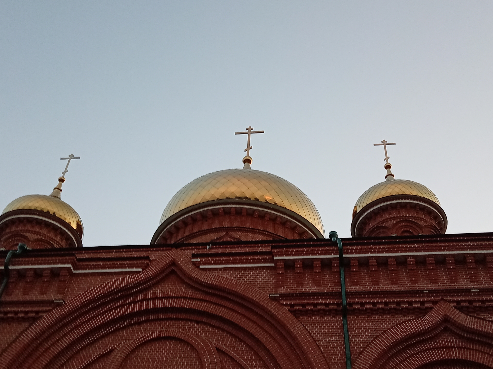 This is a low-angle, medium shot of three golden domes topped with crosses, part of a red brick church building in Savvino, Russia. The sky is clear and pale blue. The domes are the primary subjects, reflecting light and appearing smooth and rounded. The brickwork of the church is detailed, with decorative arched patterns visible. A dark green drainpipe runs vertically along the right side of the frame. There are no people or animals in the image. The overall scene is architectural and religious.
