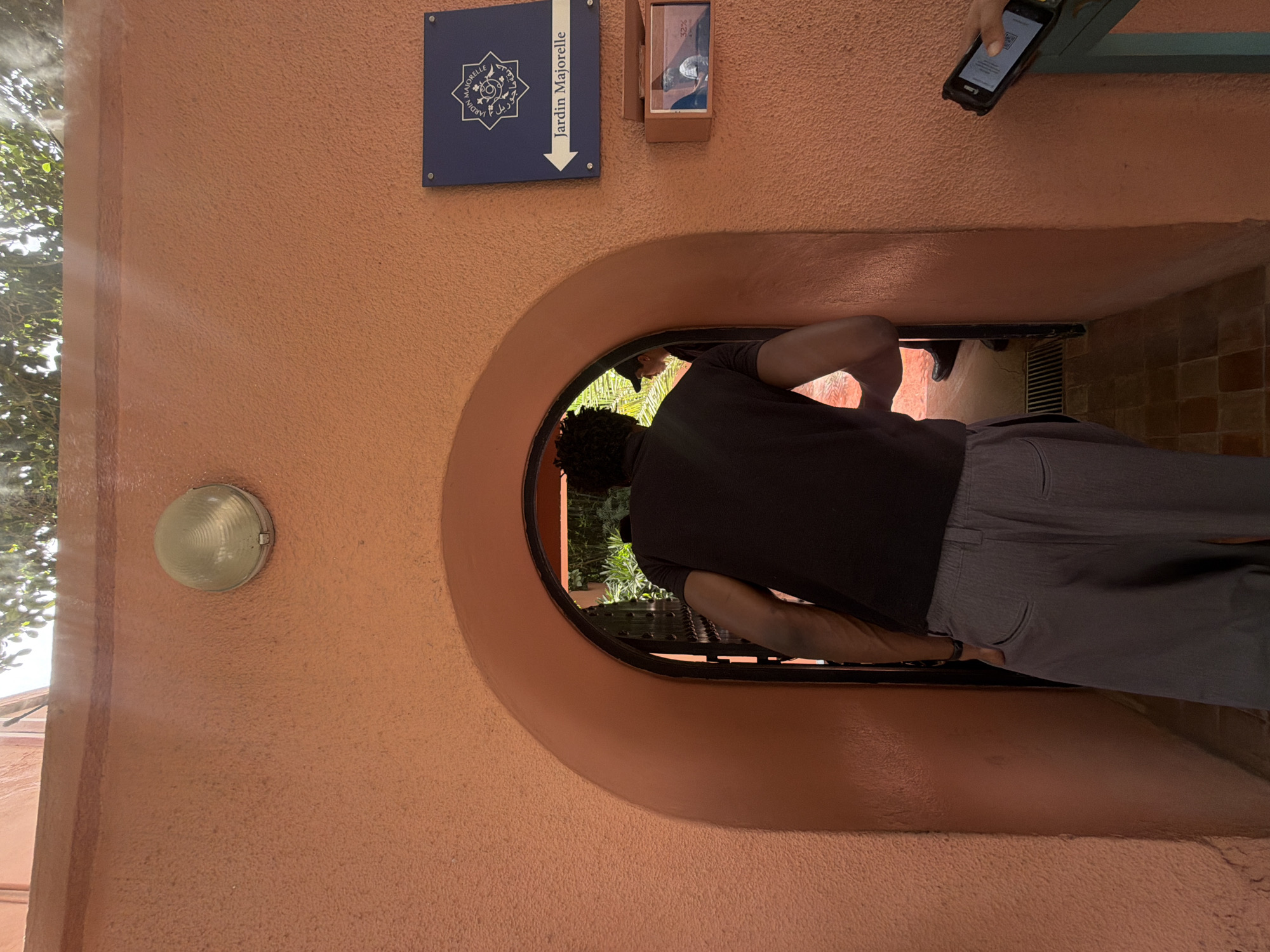 The image shows a person, viewed from behind, stooping to enter a circular opening in a peach-colored wall. The person is wearing a dark t-shirt and gray pants. The opening leads into a dimly lit space with greenery visible beyond. Above the opening, a blue sign reads "Jardin Majorelle" in white text with an arrow pointing downwards. To the right of the sign, a small brown box is mounted on the wall, containing a sliver of what appears to be a ticket or voucher with text and an image. Another person's hand is visible at the top right of the frame, holding a smartphone displaying a QR code. The scene appears to be outdoors, given the light filtering through trees on the left, suggesting it is daytime. The overall impression is of someone entering a garden attraction, possibly Jardin Majorelle in Marrakesh, Morocco, as indicated by the sign.