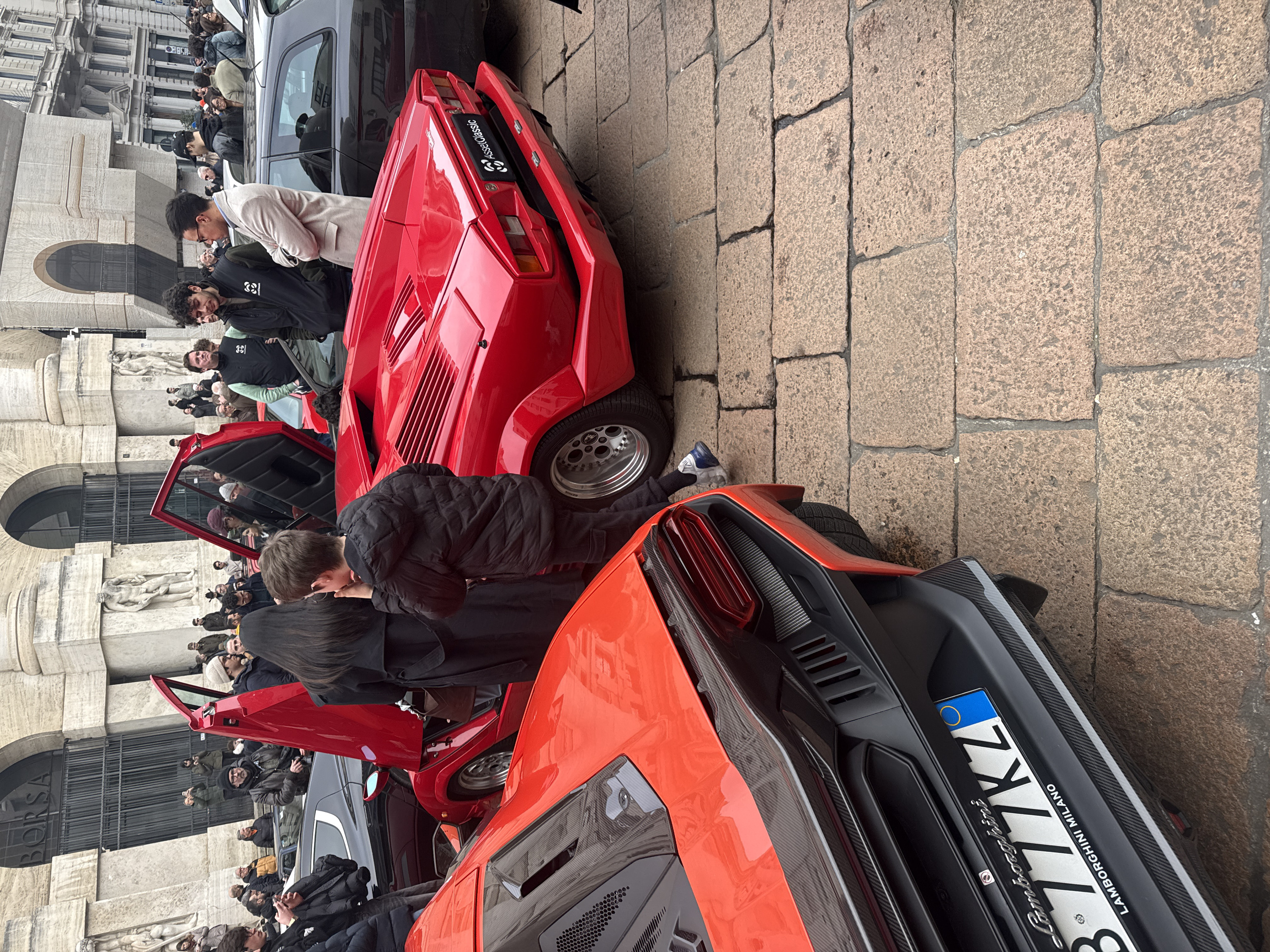 The image captures an outdoor scene on a cobblestone street in Milan, Italy, situated directly in front of the Borsa Italiana building (Palazzo Mezzanotte), identifiable by the visible "BORSA" inscription above a large arched entrance and the classical stone facade featuring prominent statues.
Two red Lamborghini supercars are central to the scene. In the midground, a red Lamborghini Countach with its distinctive scissor doors open vertically is parked. A young male, dressed in a black puffer jacket, stands beside the open driver's side door, appearing to look into the vehicle's interior. A woman in a long black coat stands partially visible next to the passenger side. The rear of this vehicle features a black plate with "AssetClassic" branding in place of a standard license plate.
In the foreground, the rear section of a more modern red Lamborghini is visible, featuring sharp lines, carbon fiber accents, and hexagonal grilles. Its Italian license plate, "B 777KZ," is prominently displayed, with "Lamborghini MILANO" printed underneath.
A crowd of approximately 30-40 individuals, comprising men and women of various ages, is gathered around the vehicles and in the background. Many individuals are observing the cars; some in the background are holding smartphones, indicating they are taking photographs or videos. People are dressed in casual, layered clothing, suggesting cooler weather. Portions of other dark-colored vehicles are visible in the background. The street is paved with irregular, light-brown cobblestones. The overall lighting is diffused, consistent with an overcast daytime sky.