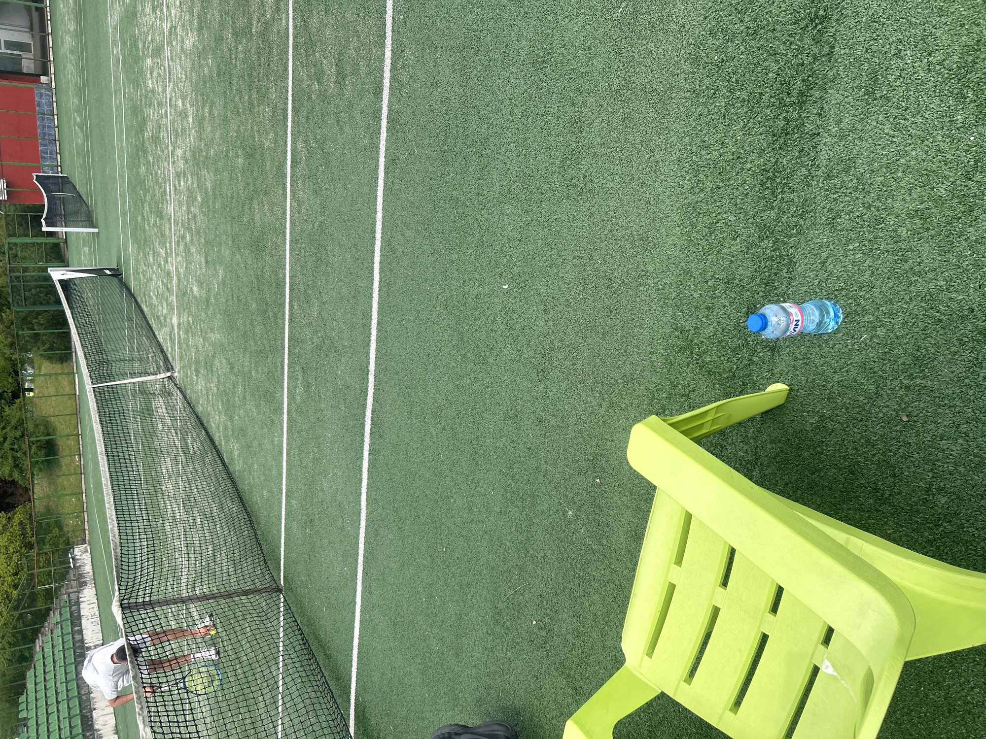 The image depicts a tennis court with artificial grass in Veliko Tarnovo, Bulgaria. A white net separates the court in half. A person wearing a white t-shirt is standing on the left side of the net. A blue water bottle and a green plastic chair are on the right side of the court. The court lines are white. The sky is overcast.