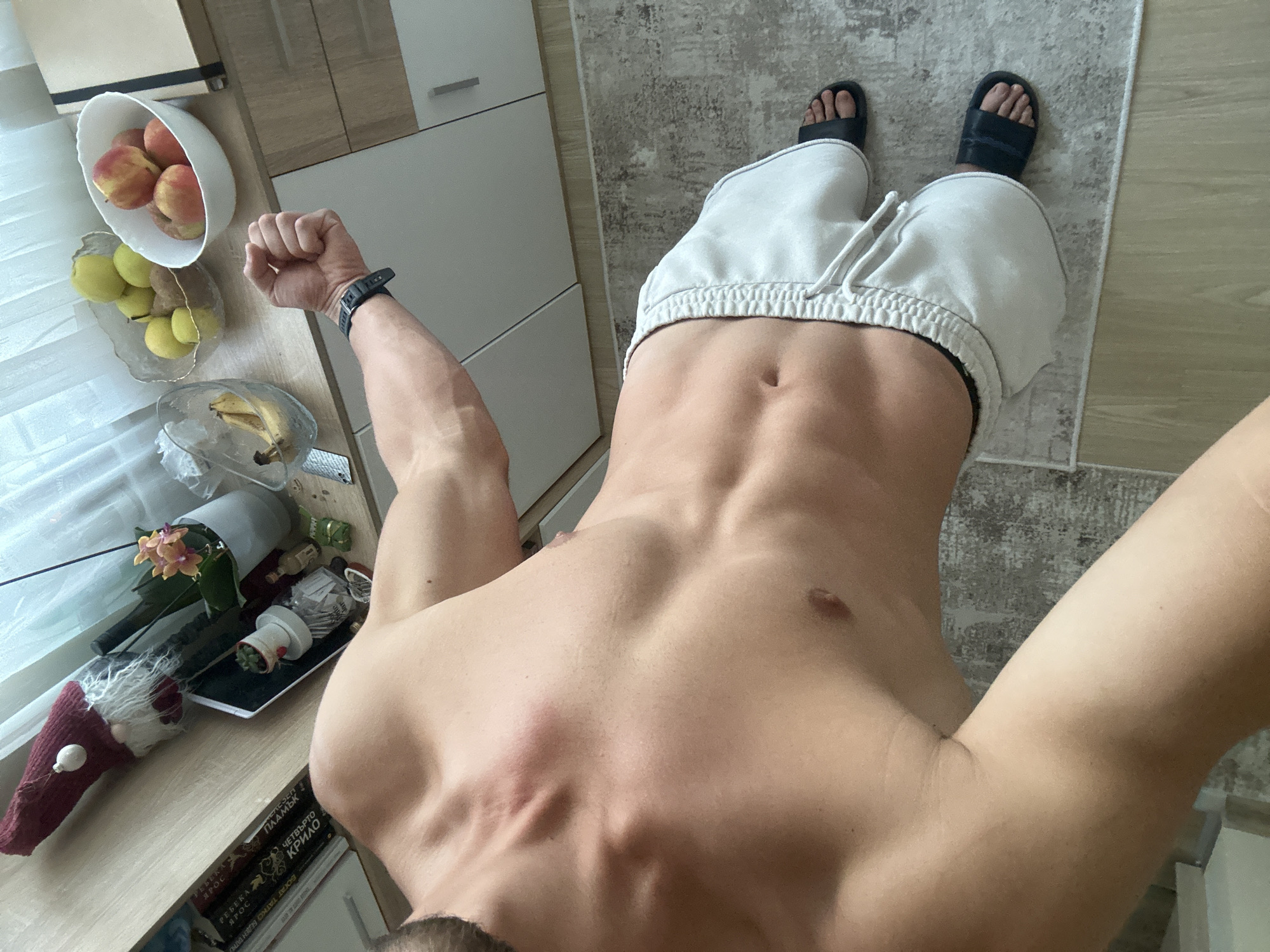 The image shows a man's torso, from mid-chest down to his feet, standing in what appears to be a kitchen or dining area. He is shirtless, wearing light-colored athletic shorts and black slide sandals. His abs are visible, and he has prominent arm veins.
To his right, on a wooden countertop, there are several bowls of fruit, including bananas, pears, and apples. Next to the fruit, there is a potted plant with orange flowers, a tablet device, and a festive gnome figurine wearing a red hat. Behind these items, a shelf is filled with books.
The man is standing on a patterned rug, and the floor around it is light-colored wood. The background includes kitchen cabinets.
The scene does not provide any specific visual cues to indicate the location as Varna, Bulgaria. The image captures a domestic indoor setting.