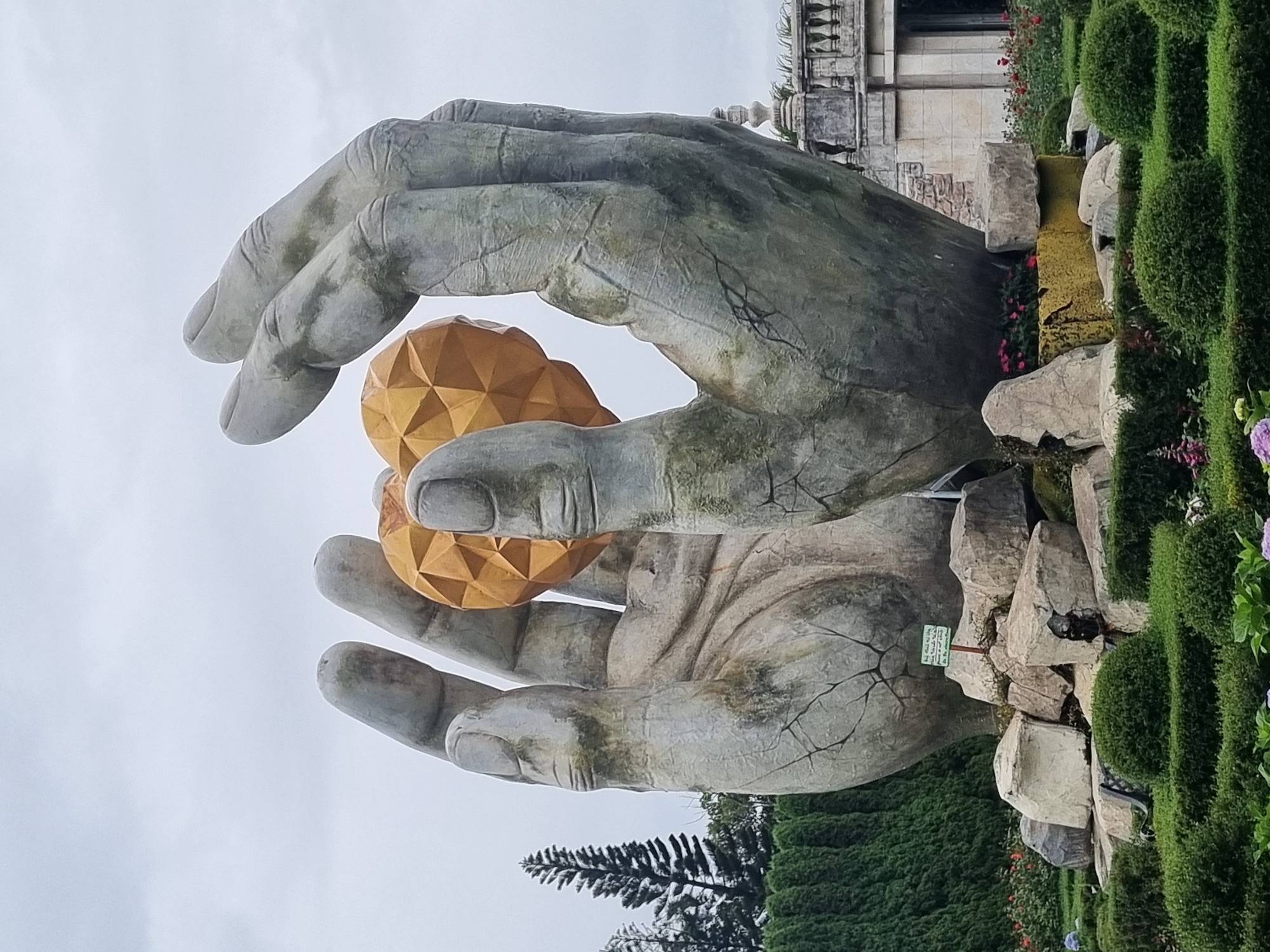 This image shows a monumental sculpture of two massive, weathered hands cradling a faceted, golden heart. The sculpture is situated outdoors, likely in a park or garden setting, with lush green hedges and scattered rocks forming the base. In the background, manicured trees and a building with classical architectural elements can be seen. The sky is overcast. This sculpture is located at the Golden Bridge, Bà Nà Hills, near Đà Nẵng, Vietnam.