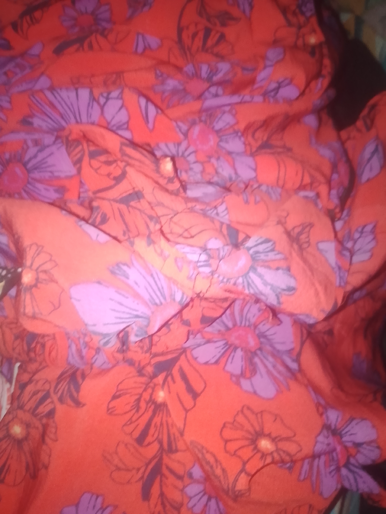 A close-up, overhead shot shows a pile of wrinkled, red fabric with a floral pattern. The flowers are large and stylized, with purple petals and dark outlines. Some of the flowers have yellow centers. The fabric is folded and draped, creating shadows and highlights. The image has a shallow depth of field, with the foreground and background being out of focus. The overall impression is one of a soft, flowing material with a vibrant and cheerful design. This image does not depict any location in the city of Jauro Yinu, Nigeria.