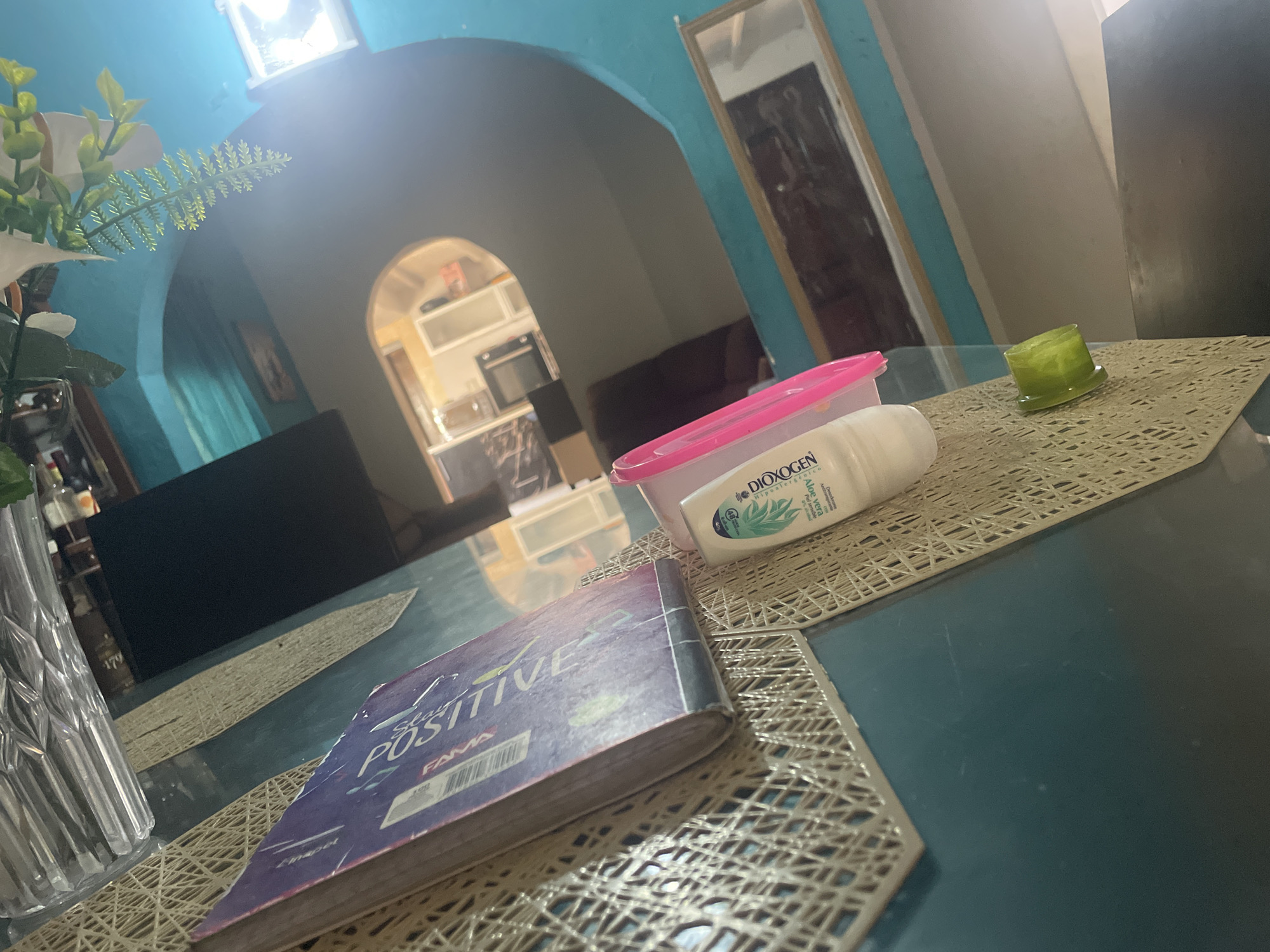 A brightly lit indoor scene, likely a living or dining area in a home in Altagracia, Venezuela, is captured from a low angle looking across a glass-top table. The room features vibrant teal-blue walls and arched doorways that reveal other parts of the house. Strong sunlight or a bright light source emanates from a rectangular window or light fixture high on the left wall, casting a lens flare and illuminating the artificial green foliage of a plant in the foreground.
On the glass table, which has several light-colored, patterned placemats, various objects are arranged. In the foreground, a purple-covered book is open, with the text "Stay POSITIVE" and "FAMA" clearly visible on its cover. Below "FAMA", smaller text includes "chapel" and "199". To the right of the book, a white roll-on deodorant with a green label and a small pink plastic container sit on a placemat. The deodorant bottle reads "DIOXOGEN" and "Aloe vera", with "HipOALERGÉNICO" beneath it, and a "OGER" logo. Further to the right, a small translucent green holder or cup is visible. A decorative clear glass vase with artificial white flowers and green leaves is partially visible on the far left of the table.
Through an arched doorway in the midground, a dark sofa or chair is discernible. Another archway in the background provides a glimpse into what appears to be a kitchen, with white cabinetry and an oven visible. A tall, rectangular mirror with a dark frame is mounted on the wall to the right. The overall impression is a casual, lived-in space, illuminated by natural light during the daytime. No people are present in the image.