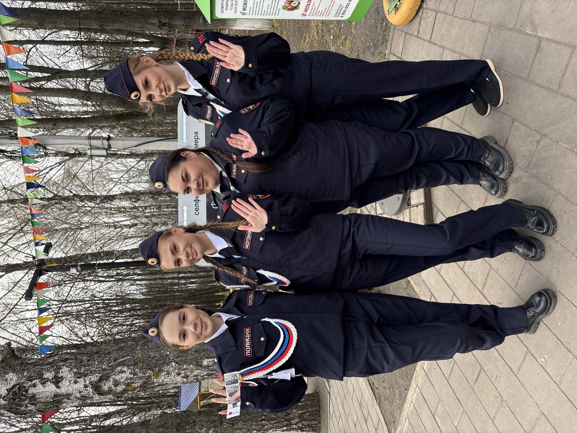 Four women in Russian police uniforms are standing in a line outdoors, posing for a photo. The first woman in line is holding a small stack of photographs in her left hand. All the women are wearing dark blue uniforms with white shirts, navy caps, and braids. The uniforms have a "полиция" patch and a red, white, and blue braided cord on the left side. The women are standing on a paved walkway. In the background, there are trees without leaves and a multicolored triangular flag banner. A sign with food-related information in Russian is visible on the right side. The city of Ulyanovsk, Russia is not mentioned in the image, and there is no indication of its location.
