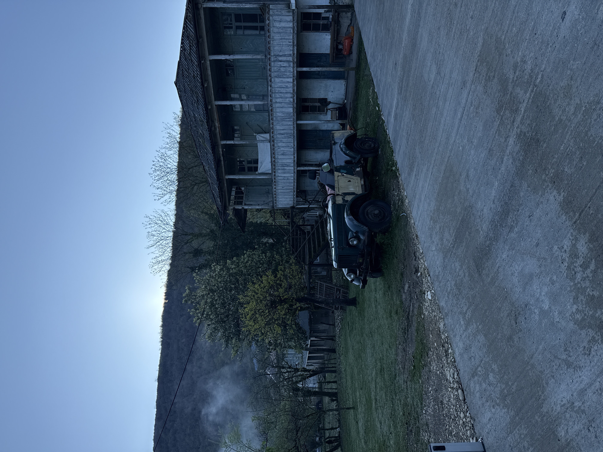 The image depicts a two-story wooden building in Gachedili, Georgia, situated next to a concrete road. A vintage tractor is parked on the grassy area in front of the building. Trees and a mountain are visible in the background, and there is smoke or mist rising in the lower left. The sky is clear and blue.