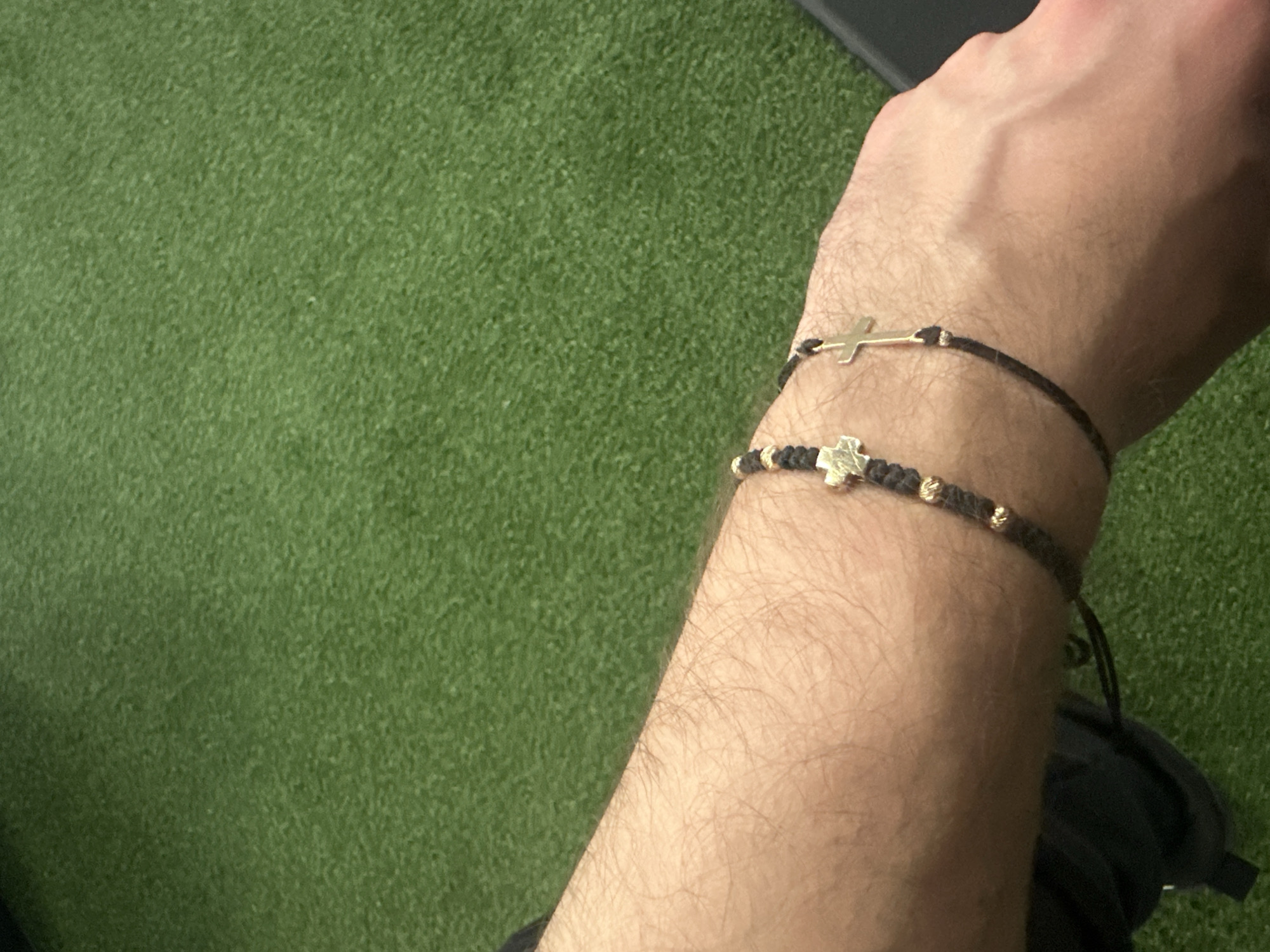 A close-up shot shows a person's forearm adorned with two bracelets. One bracelet is a thin, dark cord with a gold-colored cross. The second bracelet is thicker and features a similar dark cord interspersed with small gold-colored beads and a gold-colored clover. The person's skin is light in color, with visible arm hair. The background is a field of green. The location in the city of Plovdiv, Bulgaria cannot be determined from the image.