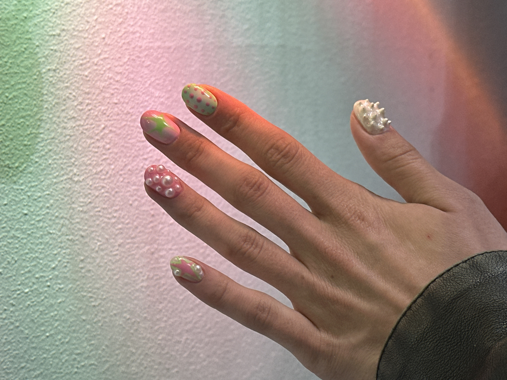 A person's left hand is shown in the foreground against a textured wall with a gradient of green and pink. The hand has various nail art designs. The index finger has a pink and green abstract design with small white beads. The middle finger has small pink pearls adhered to the nail. The ring finger has a light green base with small pink polka dots. The little finger has a white base with a raised, textured design that appears to be made of small white beads or similar embellishments. The hand is wearing a dark leather cuff on the wrist. The lighting appears to be artificial and casts warm tones on the hand.