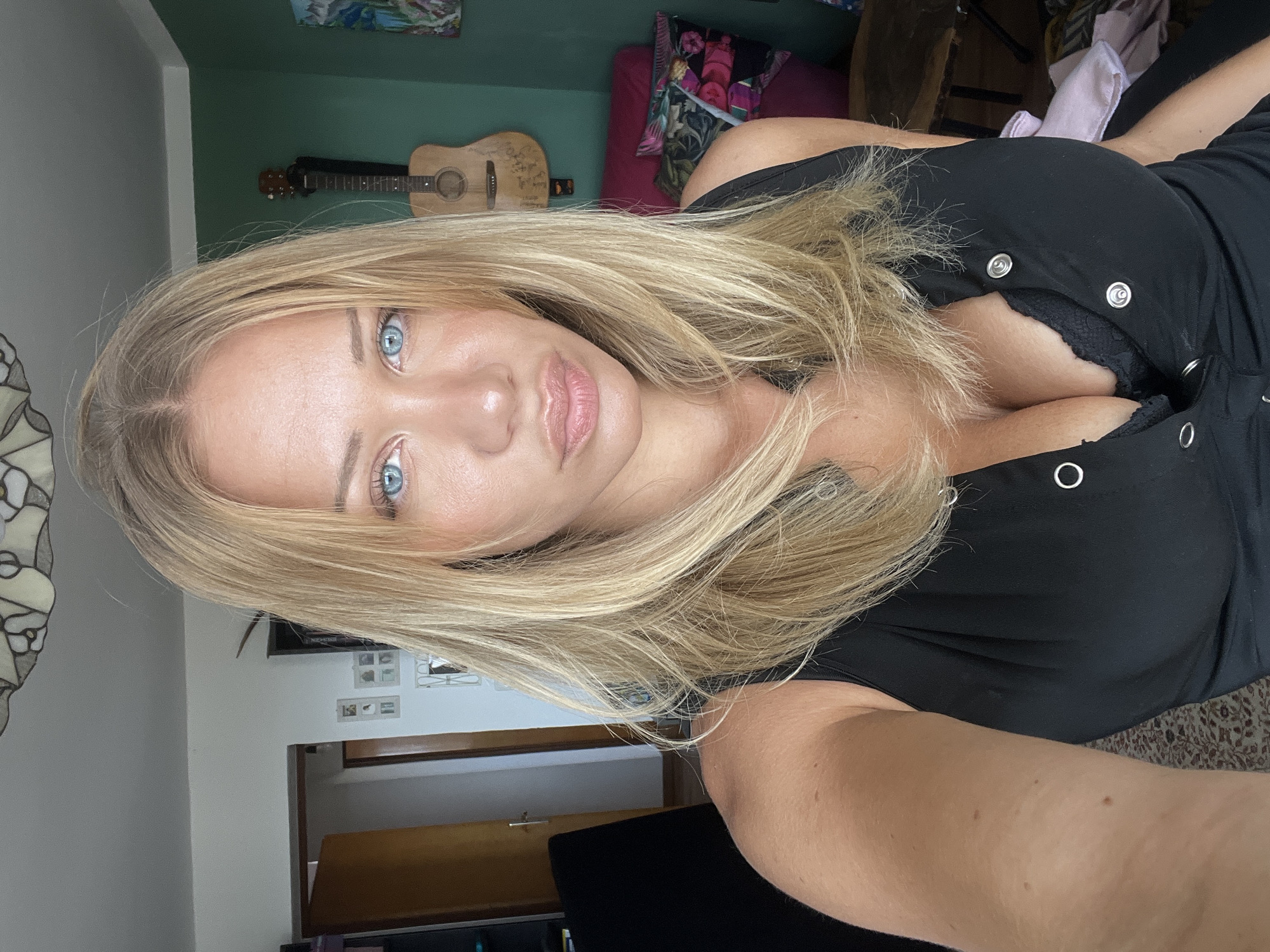 A blonde woman with bright blue eyes is taking a selfie in what appears to be an indoor residential setting. She is wearing a black V-neck top that reveals cleavage and a black bra. Her hair is long and blonde, framing her face. In the background, a guitar is hanging on a green wall, and a pink chair with decorative pillows is visible. The room also contains a doorway, a ceiling light fixture, and a rug on the floor. The lighting suggests it is daytime.