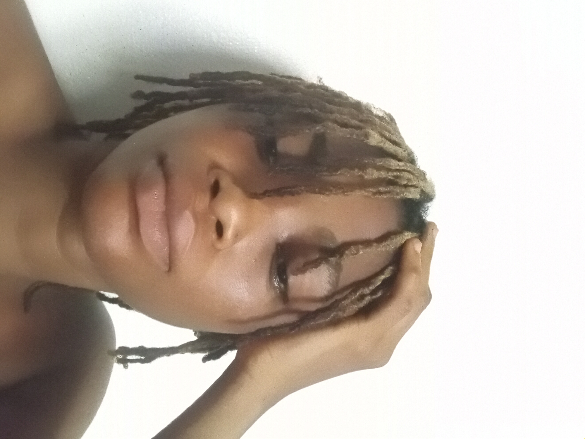 A close-up, slightly angled portrait of a brown-skinned person with dreadlocks. The person's right hand is near the right side of their face. The background is plain white. There is no indication of the location being Shagamu, Nigeria, or any other city. The person's eyes are mostly closed, and they have a relaxed expression.
