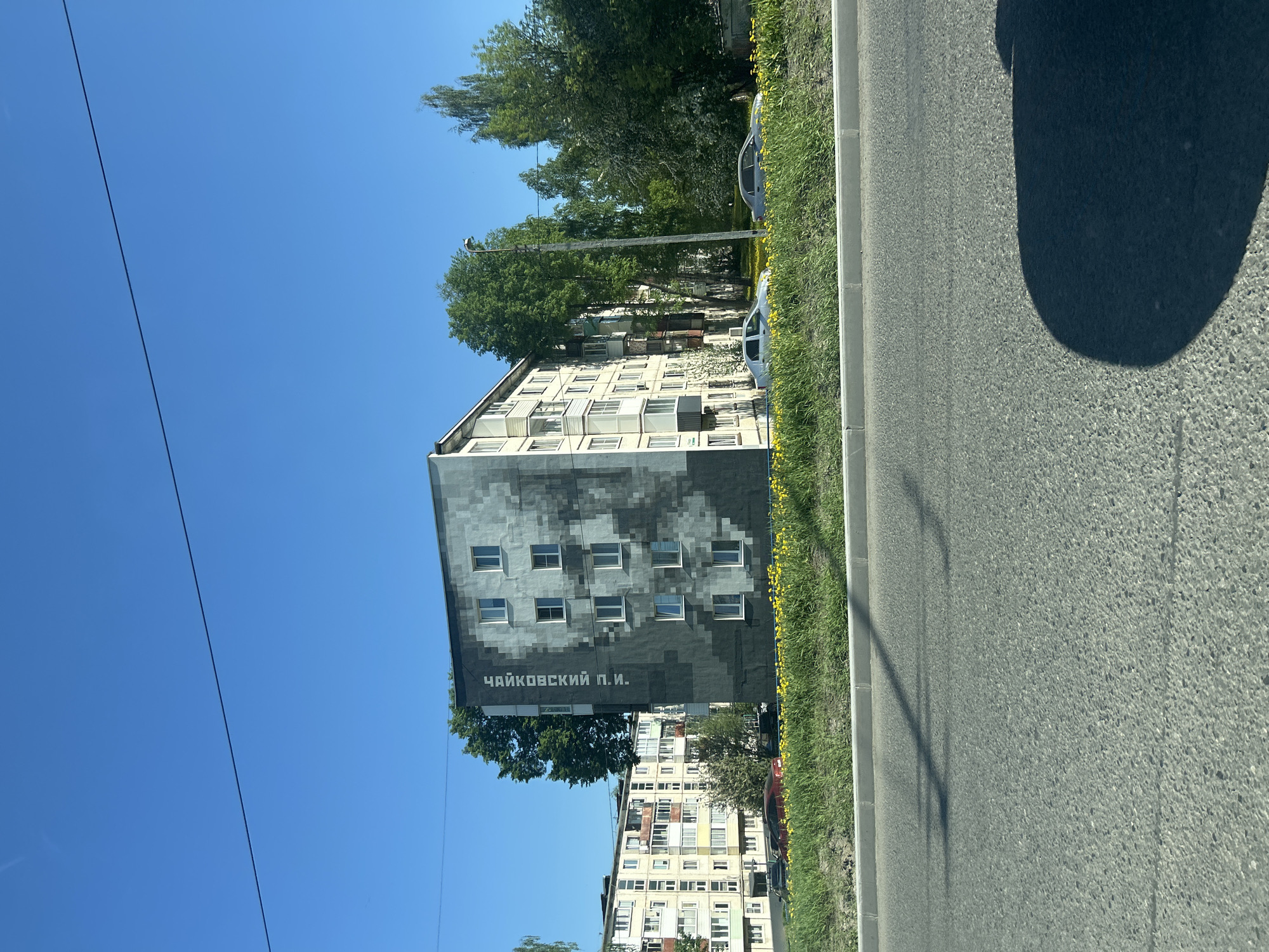 A sunny, clear-sky day photograph showcases a building in Izhevsk, Russia, prominently featuring a mural of the composer Tchaikovsky, stylized with pixelated blocks, along with his name written in Cyrillic ("Чайковский П.И."). The building is a multi-story residential apartment complex, and the mural occupies a substantial portion of its side, positioned several stories high. Adjacent to the building, green foliage and trees run along the roadside. A segment of the road and a partial shadow, likely from the vehicle from which the photo was taken, are visible. Another building is further down the road. Overhead electric lines are also visible.