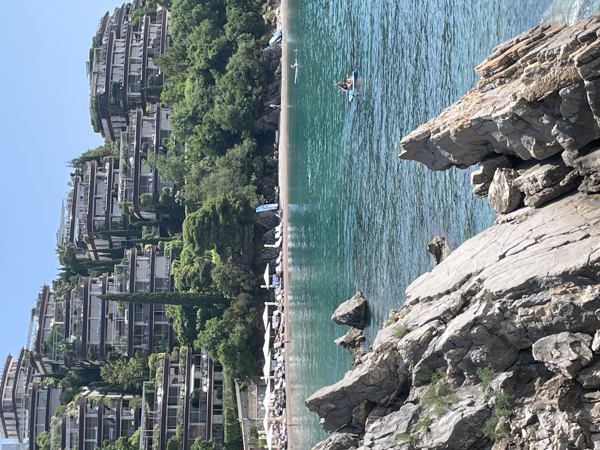 The image depicts a coastal scene, possibly in Budva, Montenegro, given the architecture. On the left, terraced residential buildings ascend a hillside, with ample greenery interspersing the structures. A beach runs along the coast, populated with white sun umbrellas and sunbathers. The water is a clear turquoise, with a person on a paddleboard visible in the distance. Rocky outcroppings are in the foreground on the right.