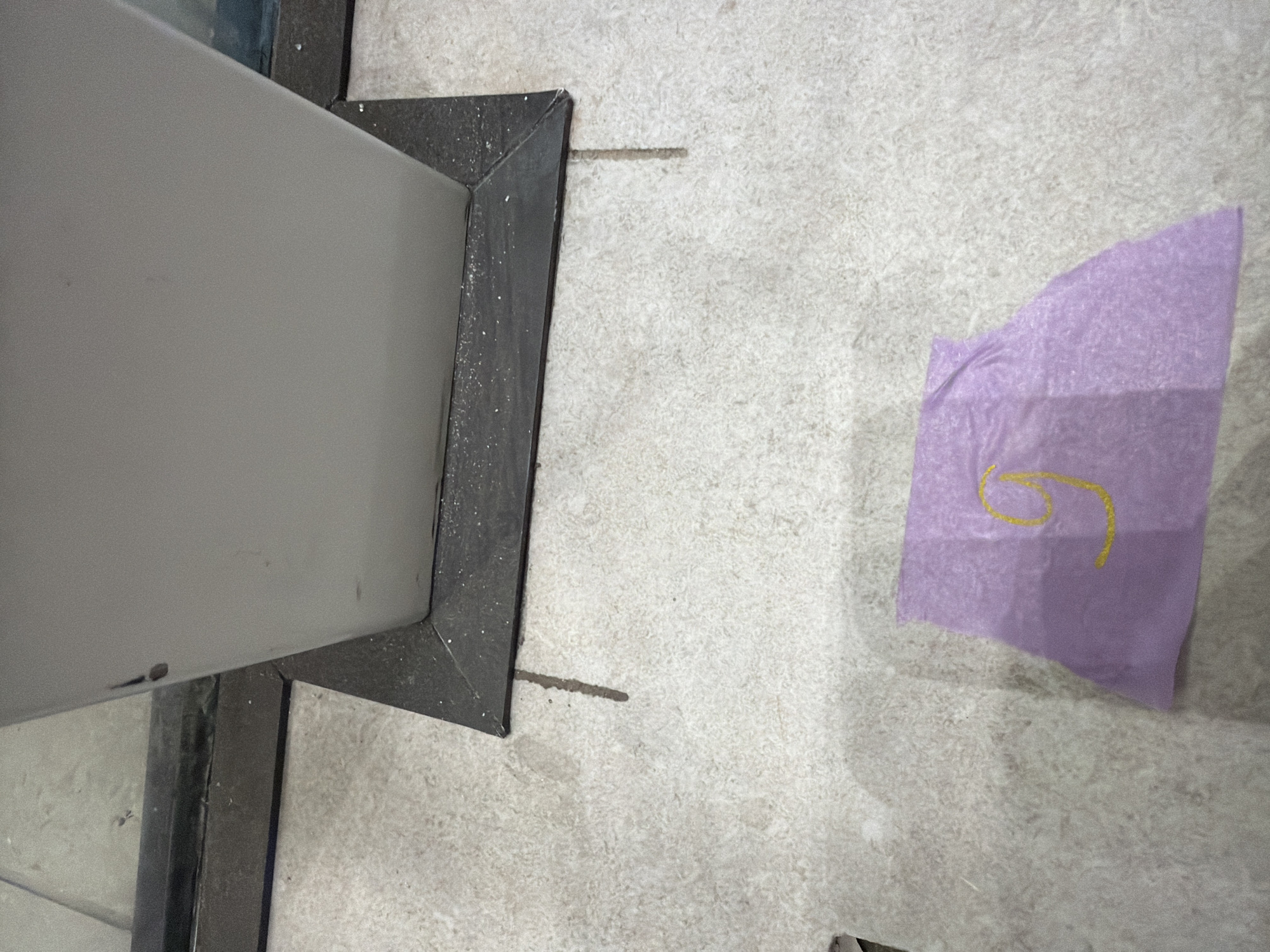 A close-up, high-angle shot shows a light gray, angular object, possibly a pillar or a display stand, situated on a textured, light-colored floor. The object has a dark gray base with a sharp, geometric edge. In the foreground, a piece of purple paper is visible, folded irregularly, with a yellow number "9" drawn on it. The floor appears to be tiled or concrete with a speckled pattern. There are some scattered white particles and dark smudges on the floor, and a dark line is visible on the floor extending from the base of the object. The background is out of focus, but suggests an indoor or sheltered outdoor environment. There are no people or animals present in the image. The image does not provide any specific indicators to pinpoint its location in Jerusalem, Israel.