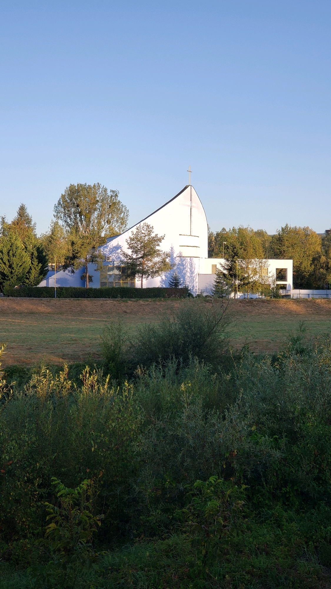 The image depicts a modern white church building with a distinctive curved roofline, topped with a cross, set against a clear blue sky. The building features large glass windows and a more rectangular annex to its right. It is surrounded by lush green trees and bushes, with a field in the foreground. The scene appears to be set in a suburban or semi-rural area. Based on the architectural style and the setting, this building is likely the Church of the Divine Providence in Prešov, Slovakia. There are no people visible in the image.