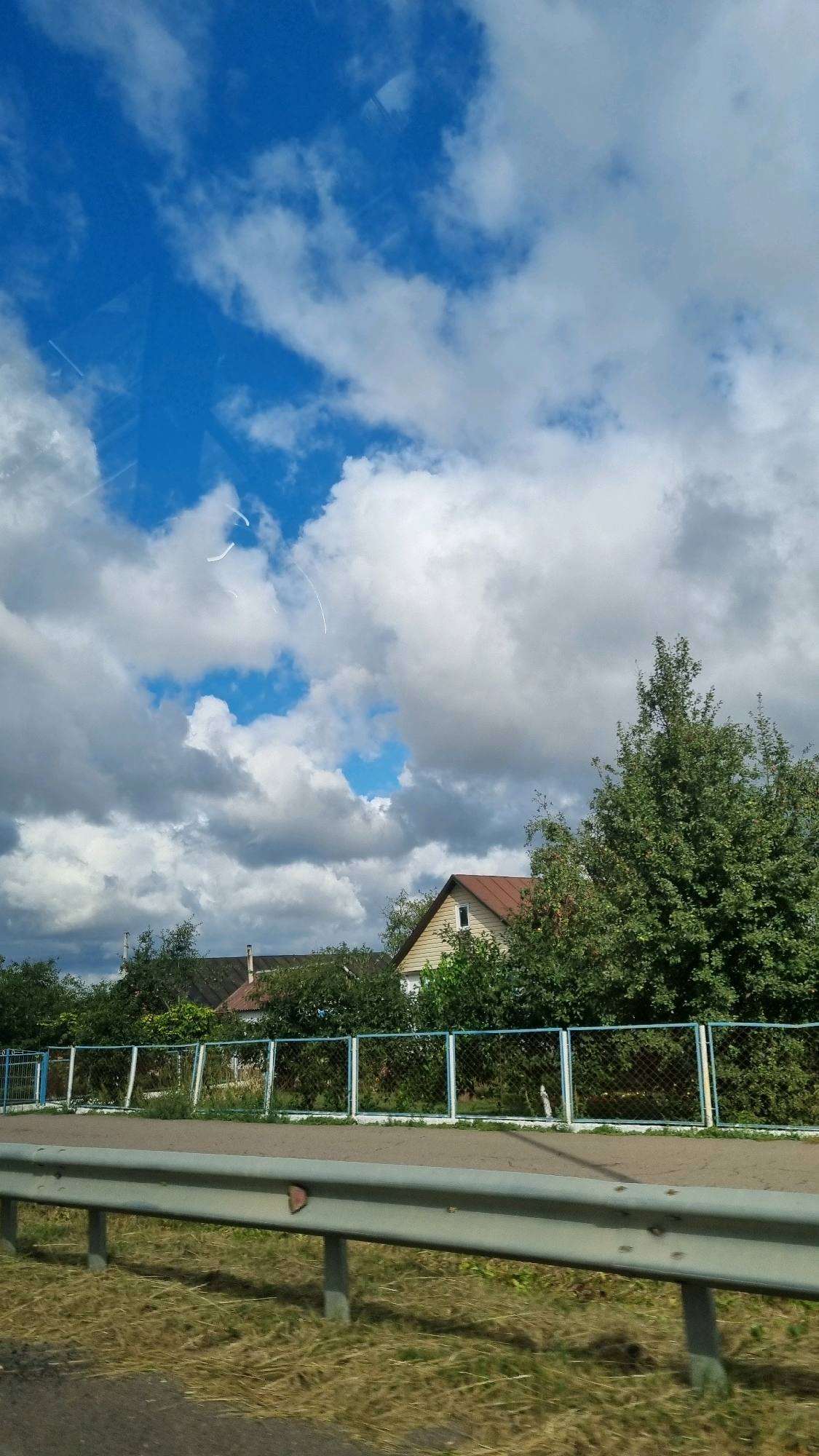 The image captures a rural scene with a blue sky filled with fluffy white clouds. The sky occupies the upper portion of the frame. Below the sky, there is a line of houses and trees. A metal fence with blue posts and a chain-link mesh separates the foreground from the background. In the foreground, a metal guardrail runs horizontally across the image, supported by concrete posts. Dry grass and a paved surface are visible beneath and around the guardrail. The overall impression is that of a sunny day in a countryside location, possibly on the outskirts of Horbakiv, Ukraine.