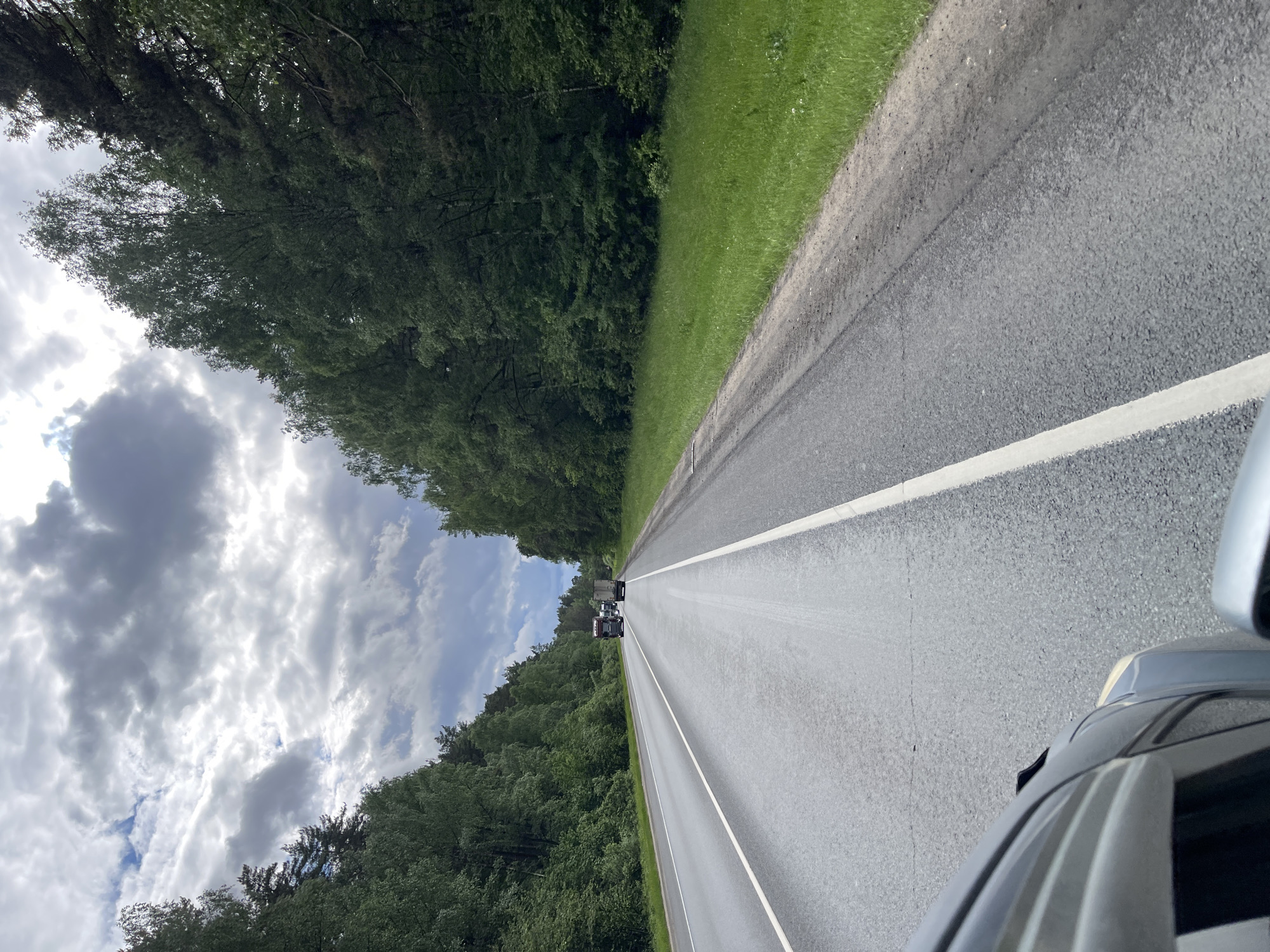 The image shows a straight asphalt road with white lines marking the lanes. Dense green trees line both sides of the road. In the distance, there are two trucks driving away. The sky above is filled with clouds. The picture appears to be taken from inside a moving car, as part of the car's roof and side mirror are visible in the lower right corner. The location is likely the road leading to or passing through Semyonovskoye, Russia given that the image was specified to be in that city.