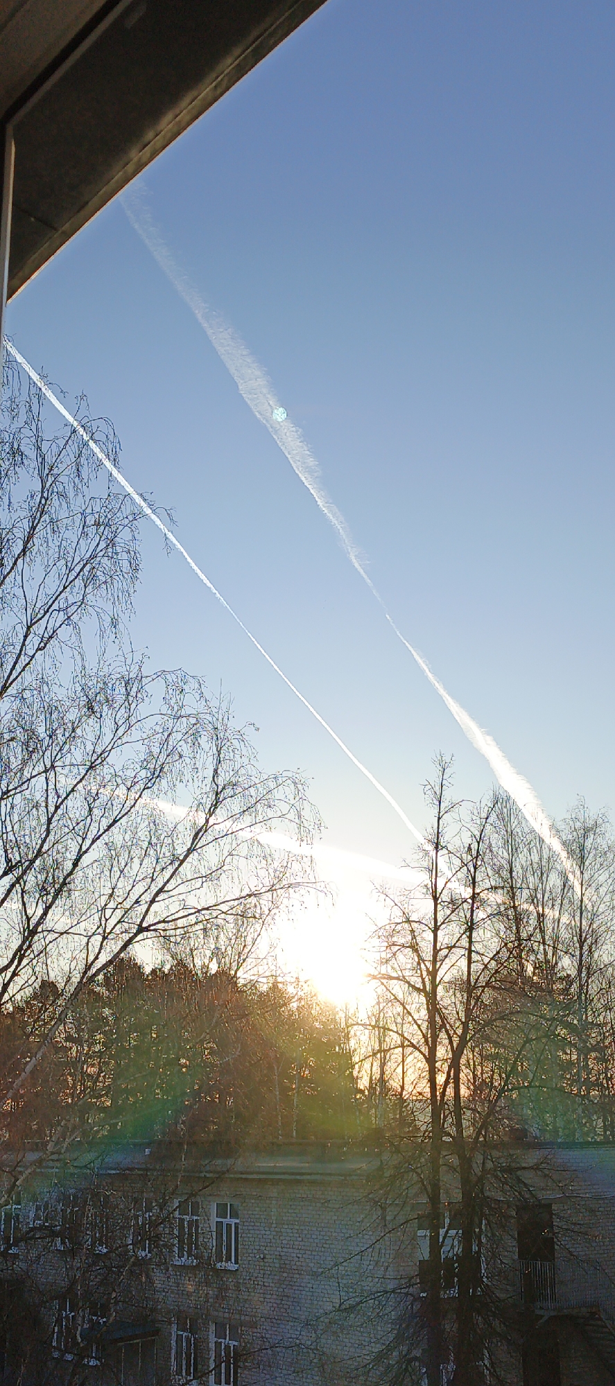 The image shows a view from a building in Chaikovsky, Russia, looking out over a landscape during sunrise. The sky is a clear blue, streaked with contrails from airplanes. In the midground, a row of buildings with windows is visible. Bare trees stand silhouetted against the rising sun, their branches intricate and delicate. The overall scene has a tranquil and serene quality, capturing a moment of natural beauty in an urban environment.