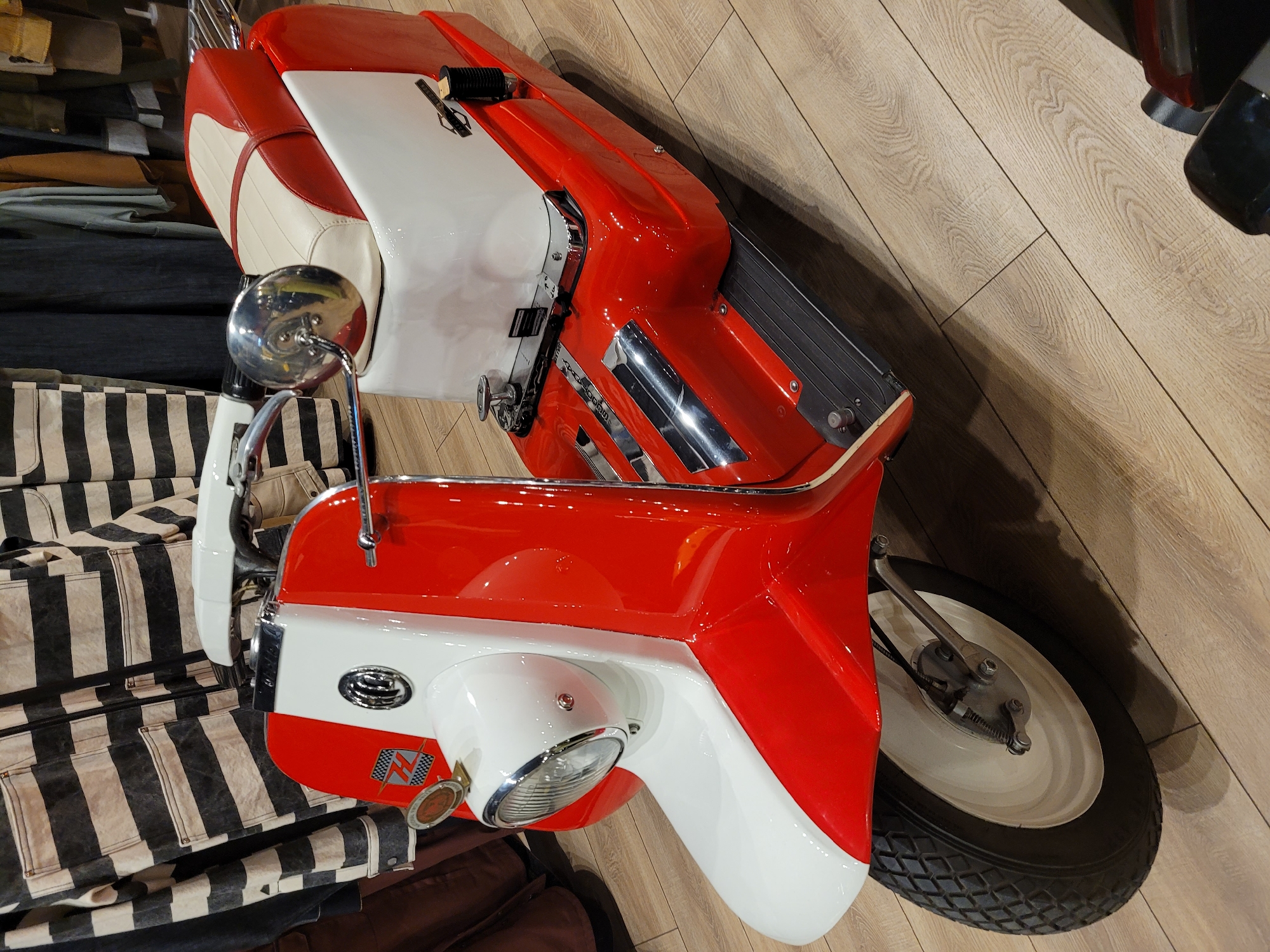 A red and white two-wheeled scooter sits on a light wood floor. It is surrounded by various fabrics and materials, including a black and white striped fabric. The scooter is parked indoors. The location is not specified in the image.