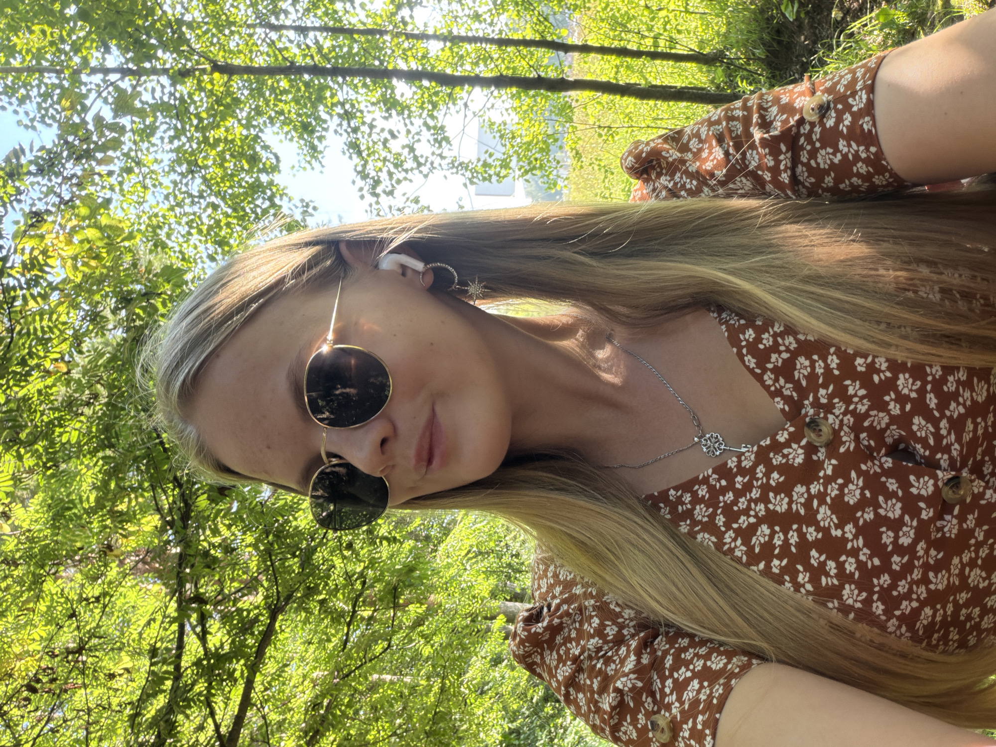 A young woman with long blonde hair is pictured outdoors, wearing sunglasses and a brown floral print dress. She has a white wireless earbud in her right ear and a silver necklace with a key pendant. The background is lush and green, suggesting a park or wooded area. There are trees and sunlight filtering through the leaves, creating a dappled effect. The image appears to be a selfie, taken from a slightly upward angle. The exact location within Izhevsk, Russia cannot be determined from the visual information alone.