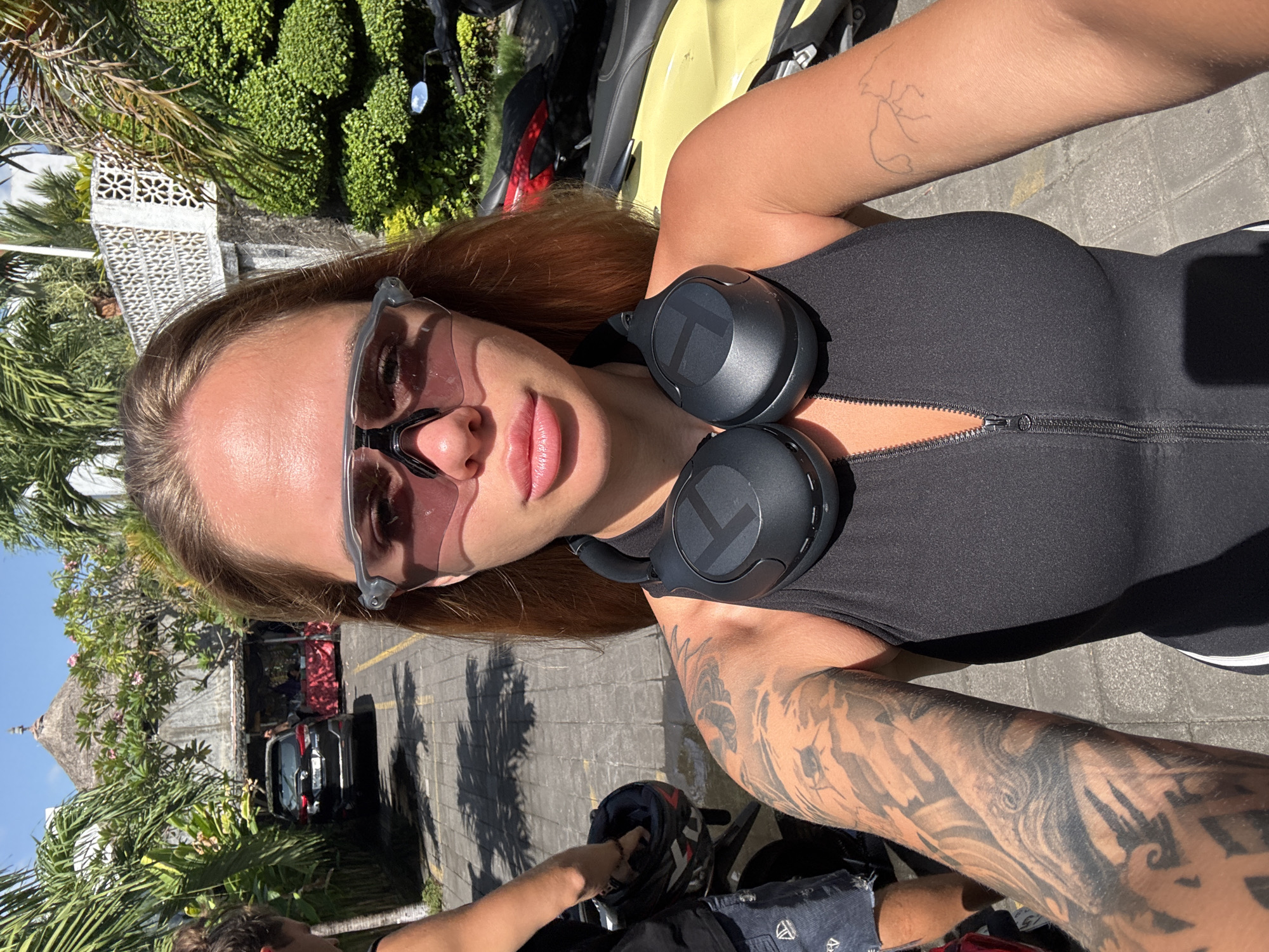 A woman with sunglasses and black headphones stands outside in North Kuta, Indonesia. She wears a black top with a zipper and has tattoos on both arms. One tattoo is a simple outline, and the other is more complex and dark. The woman is taking a selfie, partially capturing a person's arm and hand reaching into a helmet at the bottom of the frame. A yellow scooter and other vehicles are in the background, along with palm trees and a building. The ground is paved.