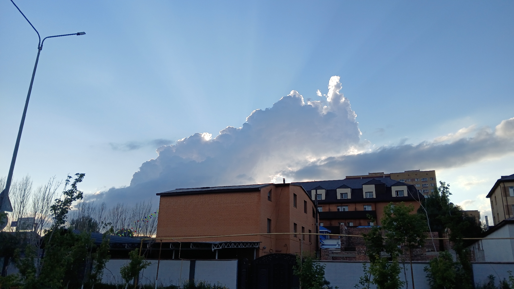 A low-angle, wide shot captures a cityscape with dramatic cloud formations against a clear blue sky. A tall, thin lamppost dominates the left side of the frame, its dark metal pole extending upwards. In the mid-ground, a two-story brick building with a dark roof and visible windows sits behind a white fence. To its right, a multi-story building with a darker roof and many windows is visible. Further right, another building with a light-colored facade and windows is partially obscured by trees. The sky features a large, bright cloud formation, backlit by the sun, creating rays of light that fan out across the sky. The lower portion of the image shows trees, fences, and the tops of smaller structures, suggesting an urban or semi-urban environment. The overall scene suggests an outdoor setting, possibly during the late afternoon or early evening, given the lighting and the dramatic cloud display.