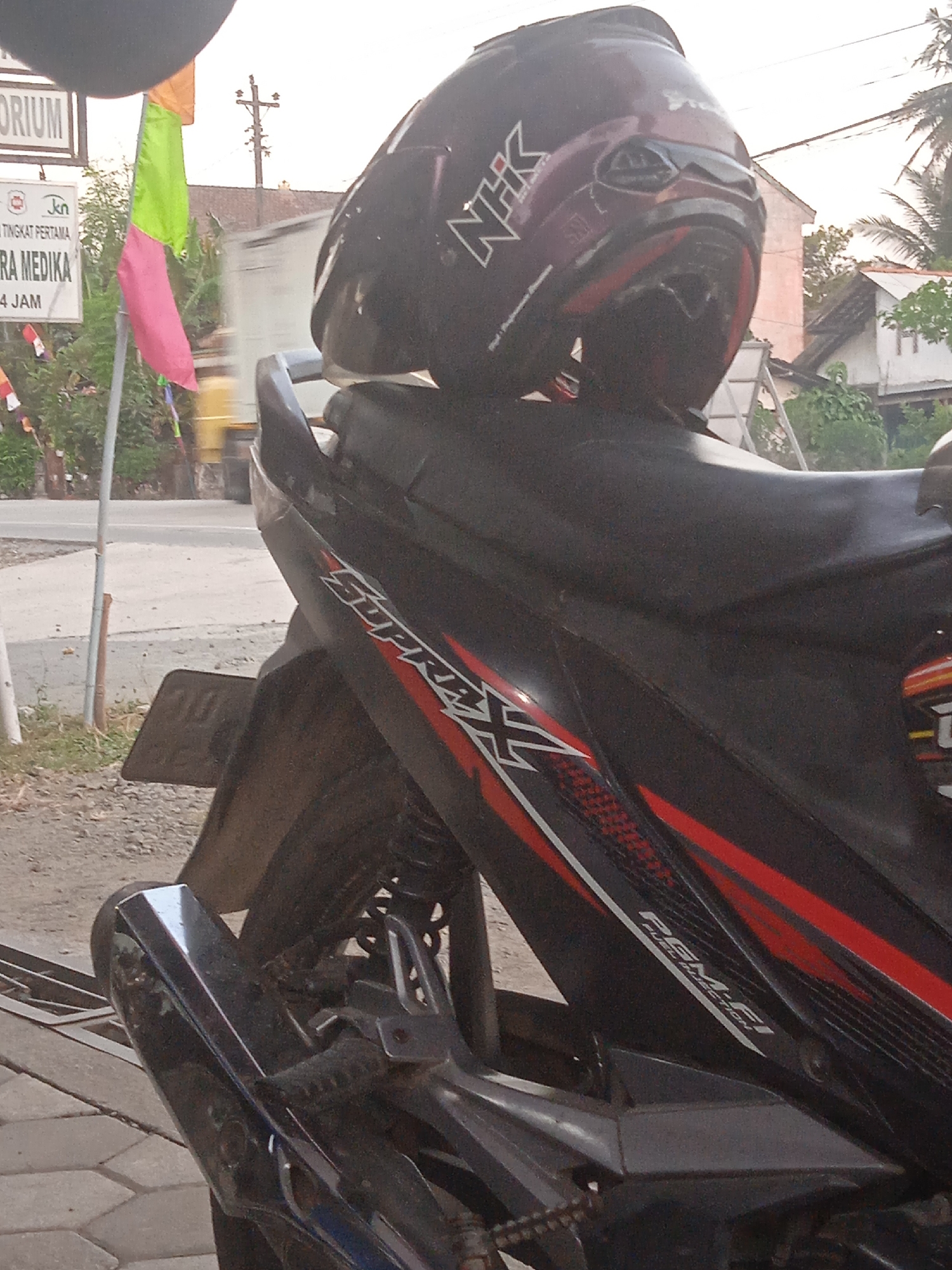 In the foreground, a black motorcycle is parked on a paved surface. A dark helmet with red accents and a white "NHK" logo is placed on the motorcycle's seat. The motorcycle's body features a "Supra X" decal in red and white, and a "PGM-FI FUEL INJECTION" decal. The rear of the motorcycle, including the exhaust pipe and license plate, is visible.
In the background, a street scene unfolds. A white sign with Indonesian text, possibly for a medical facility, is mounted on a pole. A flag with green and pink sections is attached to a pole near the sign. Further back, buildings with red tile roofs are visible, along with trees and utility poles. The sky is overcast. The overall setting appears to be a roadside in a rural or semi-urban area of Indonesia, likely Dlangu.