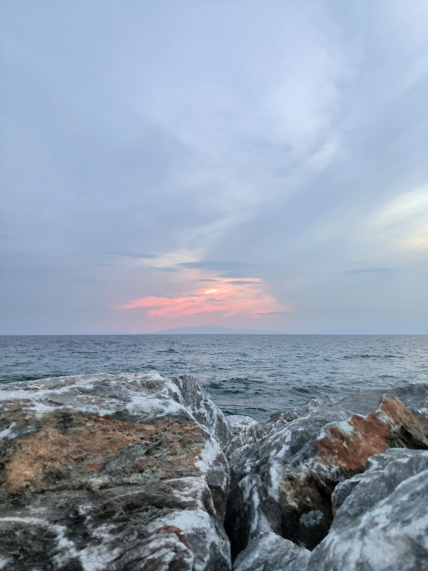 This is a vertical, eye-level shot of the sea at sunset. In the foreground, a cluster of large, jagged rocks is visible. The rocks are varied in color, with shades of gray, white, and brown. The sea stretches out to the horizon, with small waves visible. In the distance, a faint line of mountains can be seen against the horizon. The sky is filled with pale clouds, with a hint of pink and orange from the setting sun peeking through in the center. The image appears to have been taken at Nea Plagia, Greece, a coastal town known for its beaches and sunset views.
