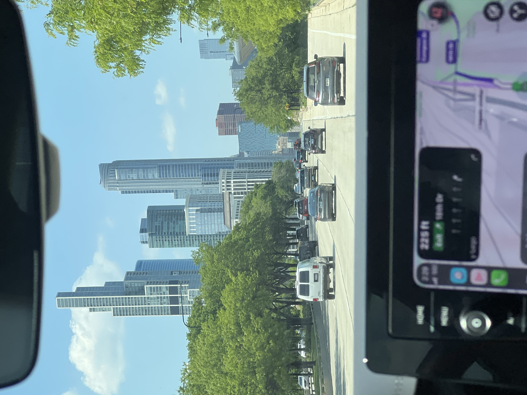 The image, taken from inside a car, shows a view of the Austin, Texas skyline. The skyline is visible in the distance, framed by trees and the road ahead, which has multiple vehicles driving on it. The car's dashboard is partially visible on the right side of the image, showing a GPS navigation display. The day appears bright and clear.