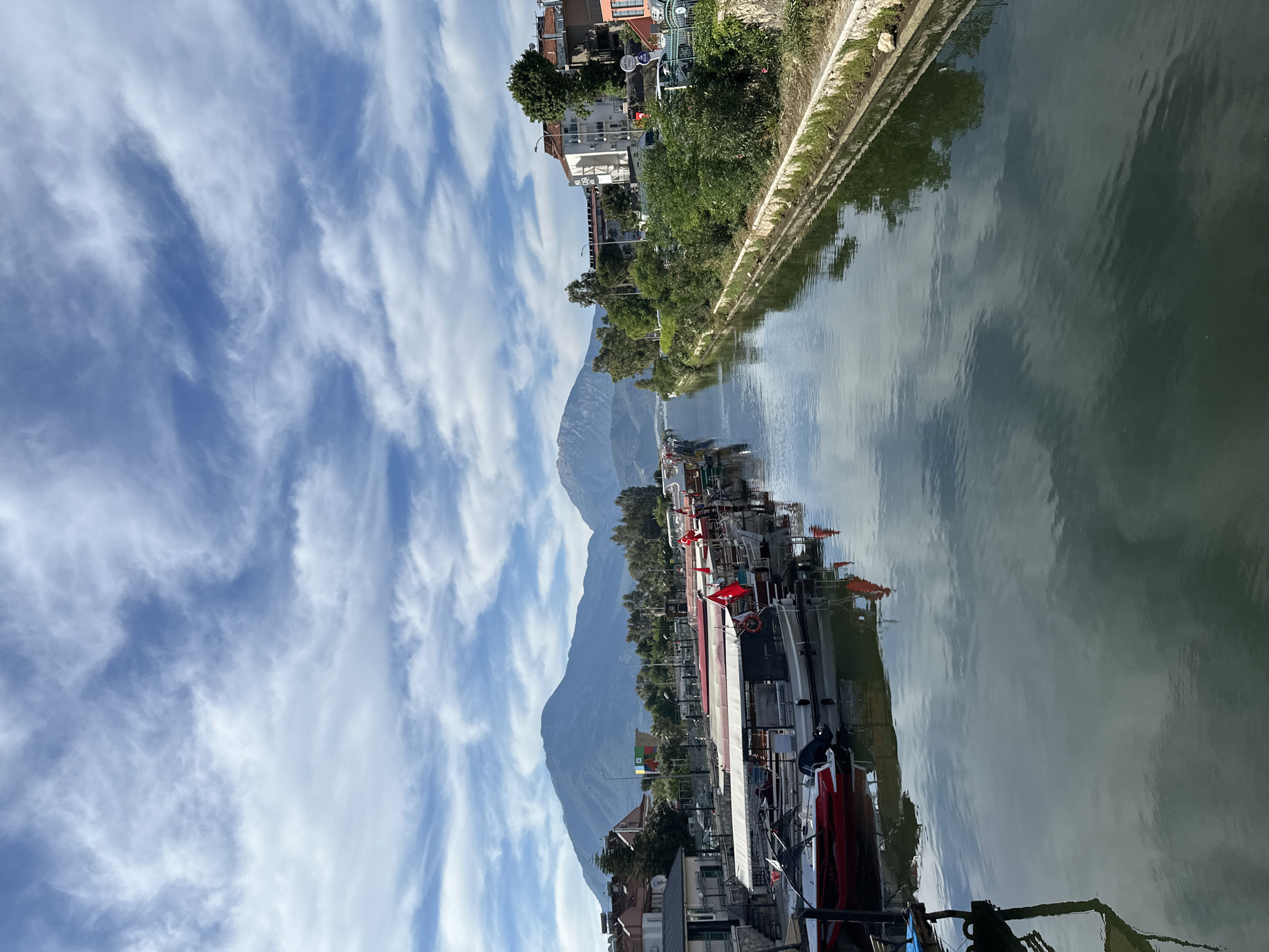 The image captures a scenic view in Fethiye, Turkey, featuring a canal bordered by buildings and lush greenery on one side and a walkway on the other. Several boats are docked along the canal, and a large mountain looms in the background, partially obscured by clouds. The sky is filled with streaky clouds, which are also reflected in the calm water of the canal.