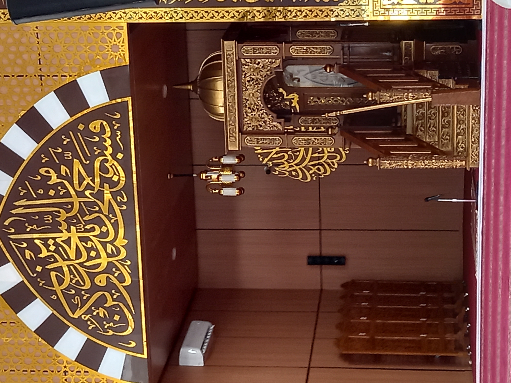 The image shows the interior of a mosque in Samarinda, Indonesia, specifically focusing on the ornate mimbar (pulpit). The mimbar is constructed of dark wood with intricate gold carvings and a golden dome at the top. A staircase with carved railings leads up to the pulpit. To the left of the mimbar, there is a large golden Arabic calligraphy inscription mounted on the wall. Above the mimbar, an arched structure with a decorative geometric pattern and alternating brown and white stripes is visible. Hanging from the ceiling is a chandelier with multiple light fixtures. In the background, a white air conditioning unit is mounted on a wooden paneled wall. Wooden room dividers are also present. The floor is covered with red carpet. A microphone on a stand is positioned in front of the mimbar.
