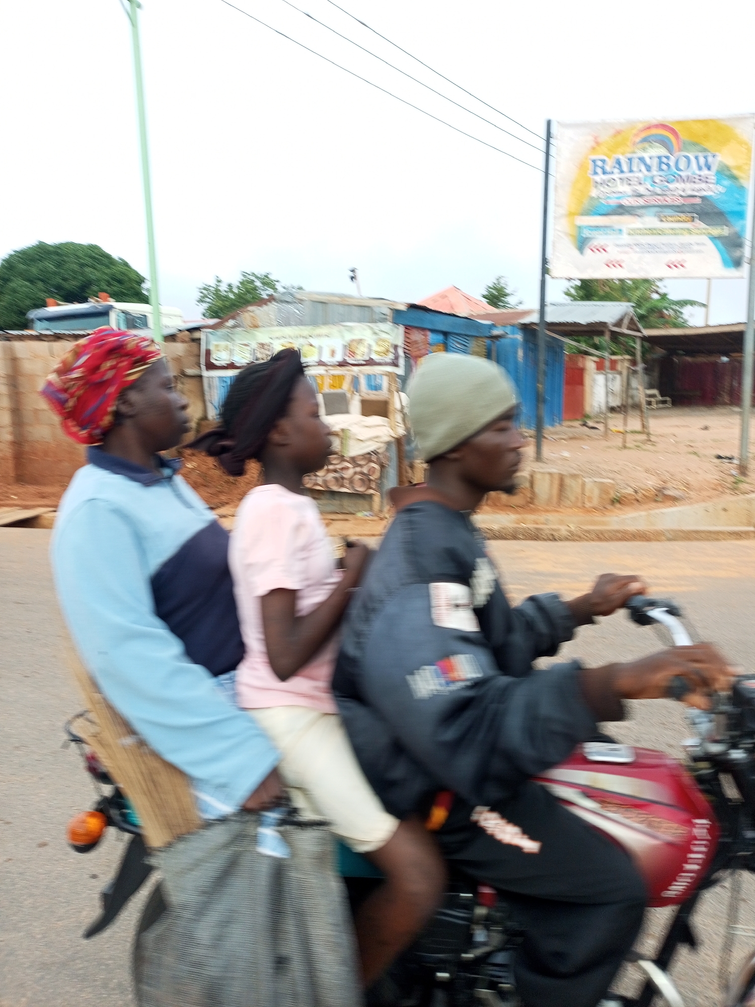 In Garko, Nigeria, three individuals are captured in motion on a motorcycle. The rider, a man wearing a grey beanie and a dark jacket, is at the helm. Behind him, a woman with a red headscarf and a blue and dark blue long-sleeved shirt is seated. A young girl, with a dark headscarf and a pink t-shirt, is positioned behind the woman. The motorcycle has a red fuel tank and is equipped with a basket-like object attached to the rear. The background features a dusty, unpaved road bordered by modest buildings, including one with a prominent "RAINBOW HOTEL GOMBE" sign. Overhead, power lines are visible against a cloudy sky. The image is blurry, indicating movement, likely from the motorcycle in transit.