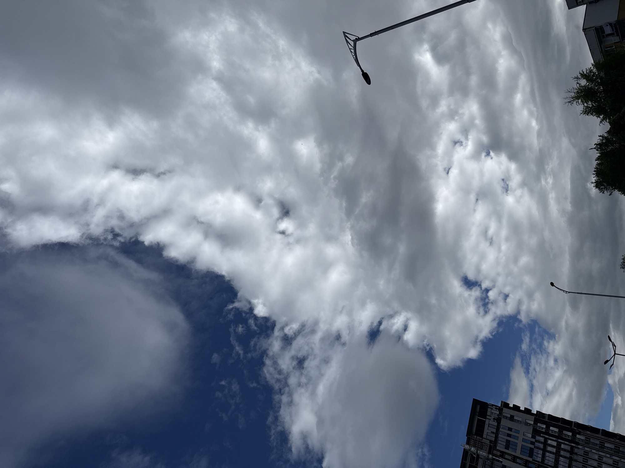 The image shows a low-angle shot of the sky in Plovdiv, Bulgaria, revealing a mix of dark blue clear sky and thick white and gray clouds. A modern, high-rise building with multiple windows and a dark exterior dominates the lower right corner. Two streetlights extend into the frame from above and the right, with visible curved arms and spherical bulbs. Green foliage from trees is visible at the top right corner.