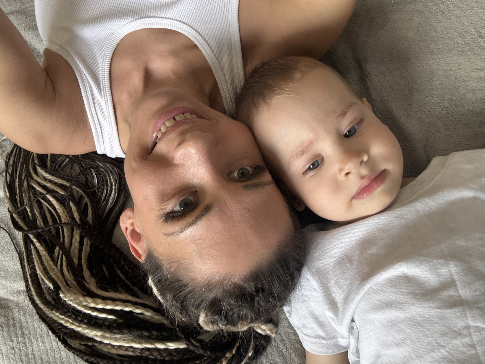 A woman and a young child are lying on a gray surface, presumably a bed or couch. The woman has tan skin, brown eyes, and braided dark and light hair. She is wearing a white tank top. The child has fair skin, light blue eyes, and short light brown hair. They are wearing a white t-shirt. The woman is smiling, and the child is looking at the camera. The location of the image is not specified.