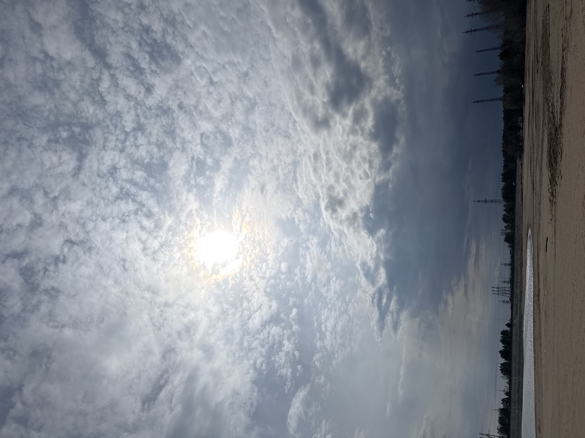 The image shows a view upwards towards a bright sun obscured by a sky filled with altocumulus clouds. The horizon line, tilted to the right, reveals a landscape with power lines and trees against a sky with varying cloud cover. A long, narrow body of water borders the landscape. The scene is likely in Noviy, Russia, based on the prompt.