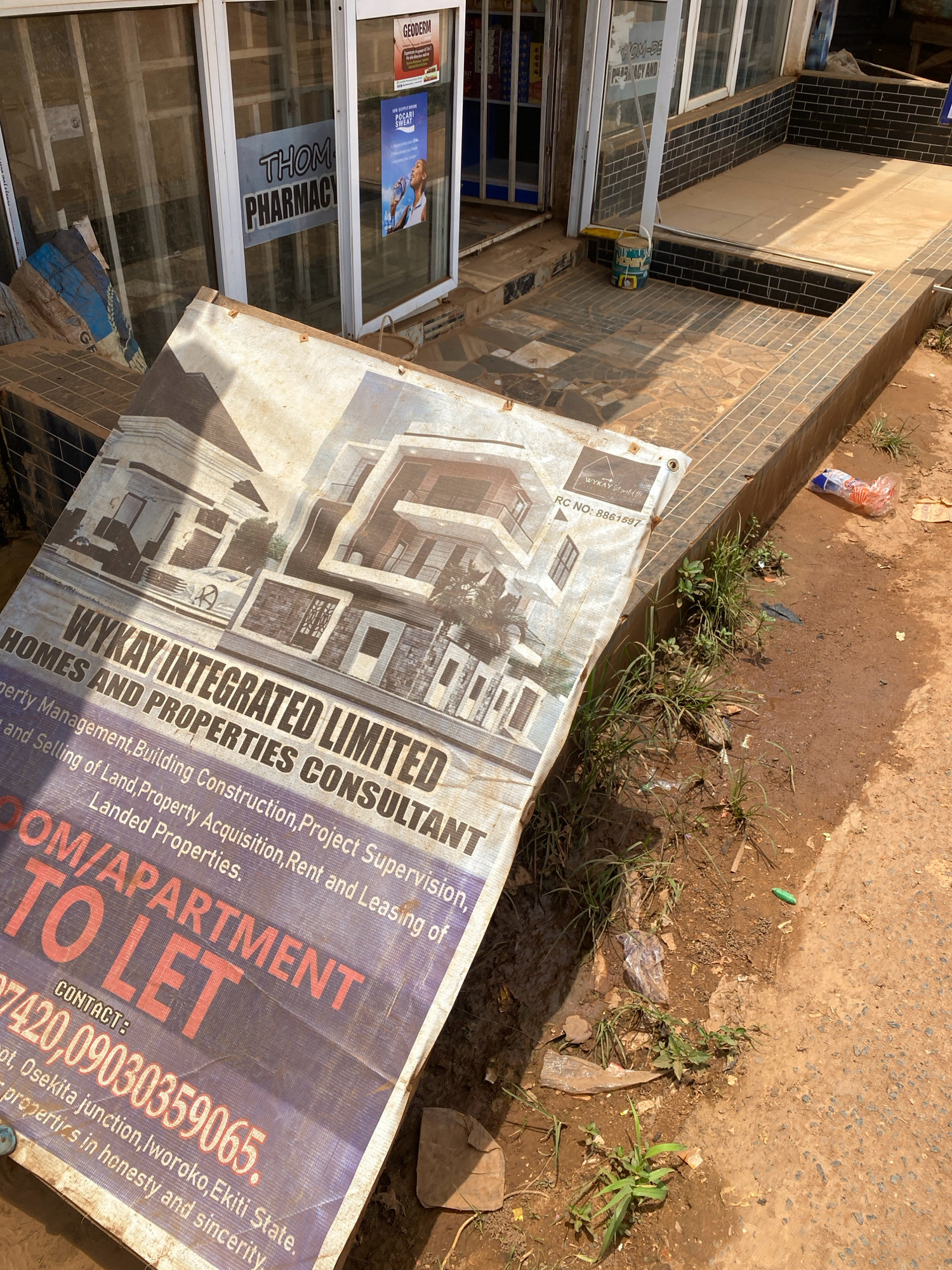 The image shows a close-up of a tattered, off-white banner advertising "WYKAY INTEGRATED LIMITED HOMES AND PROPERTIES CONSULTANT". The banner is draped over a tiled step in front of a building that appears to be a pharmacy, with signage for "THOM PHARMACY" visible. The banner details services such as "Property Management, Building Construction, Project Supervision, and Selling of Land, Property Acquisition, Rent and Leasing of Landed Properties." A section of the banner displays "ROOM/APARTMENT TO LET" in large, red lettering, followed by a contact number: "07420,09030359065". The location is identified as "Ot, Osekita junction, Iworoko, Ekiti State."
The environment appears to be an outdoor commercial area. The ground is a mixture of reddish-brown soil and cracked paving stones. Greenery, consisting of grass and small plants, is growing in patches along the edge of the paved area and the building. There is some litter scattered on the ground, including plastic bags and a discarded orange and clear plastic bottle. The building's storefront has large glass windows and a tiled facade. Sunlight casts shadows, indicating it is daytime. The overall impression is of a somewhat neglected commercial street in Iworoko, Nigeria, with a property rental advertisement prominently displayed.