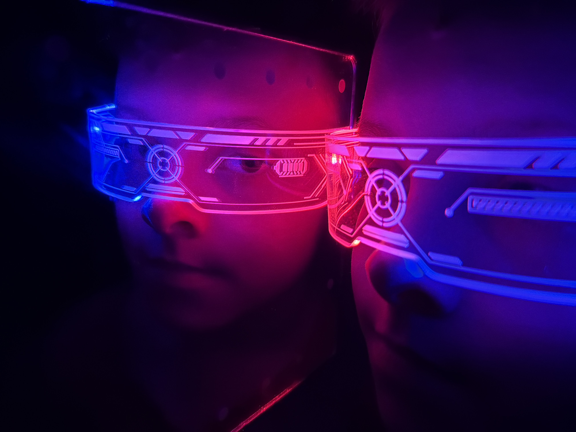 Two people wearing illuminated, transparent, futuristic glasses are depicted. The glasses on the left person are illuminated with a blue light, while the glasses on the right are illuminated with a red light. The background is dark. There is no evidence to suggest the photo was taken in Votkinsk, Russia.