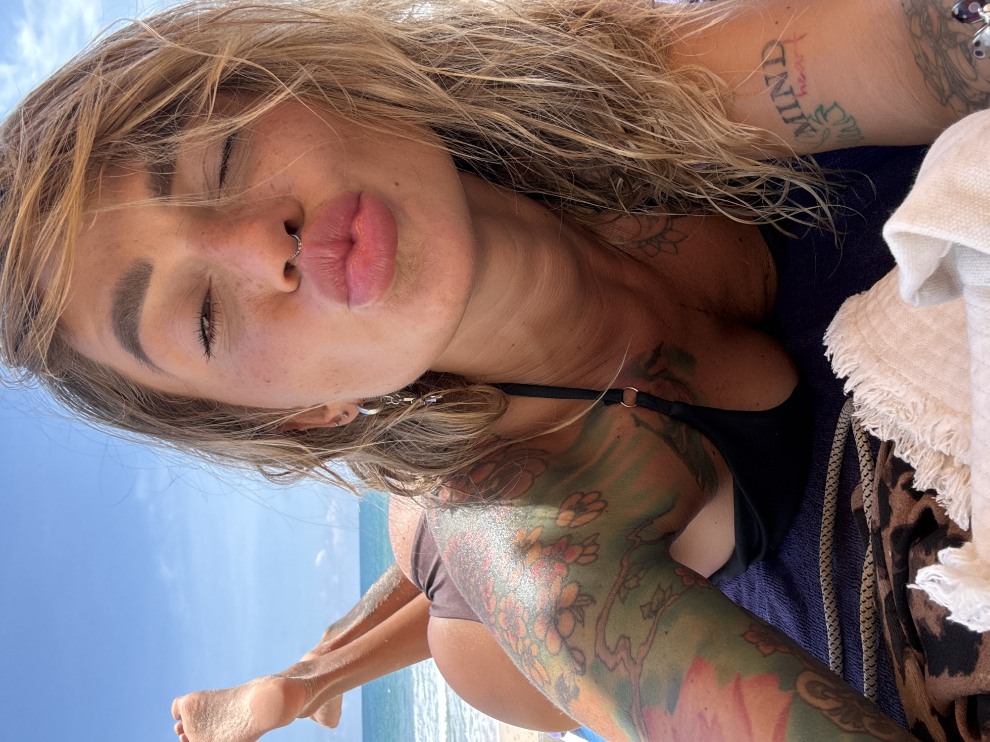 A woman with blonde hair, freckles, and tattoos on her arm and shoulder is lying on a towel on a beach in Choeng Thale, Thailand. She is wearing a brown bikini top and has her eyes closed while puckering her lips. The ocean and a clear blue sky are visible in the background.