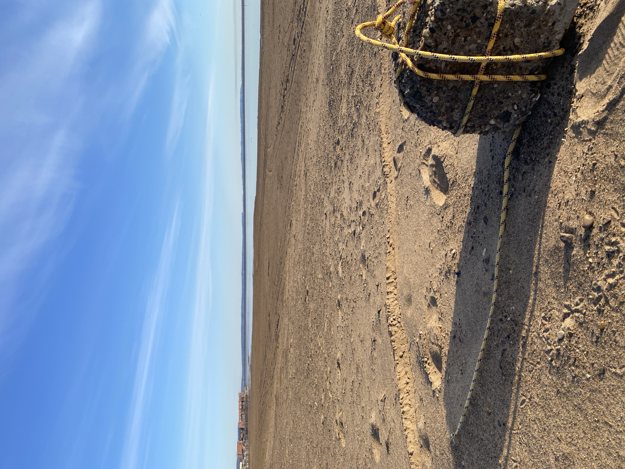 The image presents a wide, expansive view of a sandy and gravelly shore next to a large body of water, characteristic of a river or reservoir landscape, located near Chaikovsky, Russia. The scene is bathed in clear, bright daylight under a vibrant blue sky, adorned with scattered, thin streaks of white clouds that gradually fade to a lighter shade towards the horizon.
In the immediate foreground, positioned to the right, is a substantial, rough-textured concrete block or large rock. A distinctive yellow rope with a black pattern is wrapped around this block, with a segment trailing across the textured ground, suggesting it might be an anchor or marker. Long, distinct shadows are cast across the sand, indicating a low sun angle, likely early morning or late afternoon.
The ground itself is marked by visible tire tracks or vehicle imprints, alongside several human footprints, one of which is clearly defined in the sandy foreground. The extensive shore stretches towards a calm, light blue body of water that occupies the middle distance. Along the far horizon, a narrow strip of distant land is visible, featuring small, indistinct buildings or structures, suggesting a settlement on the opposite bank. No people are visible, and no text is present in the image. The overall impression is one of a tranquil, open natural environment subtly marked by human activity.