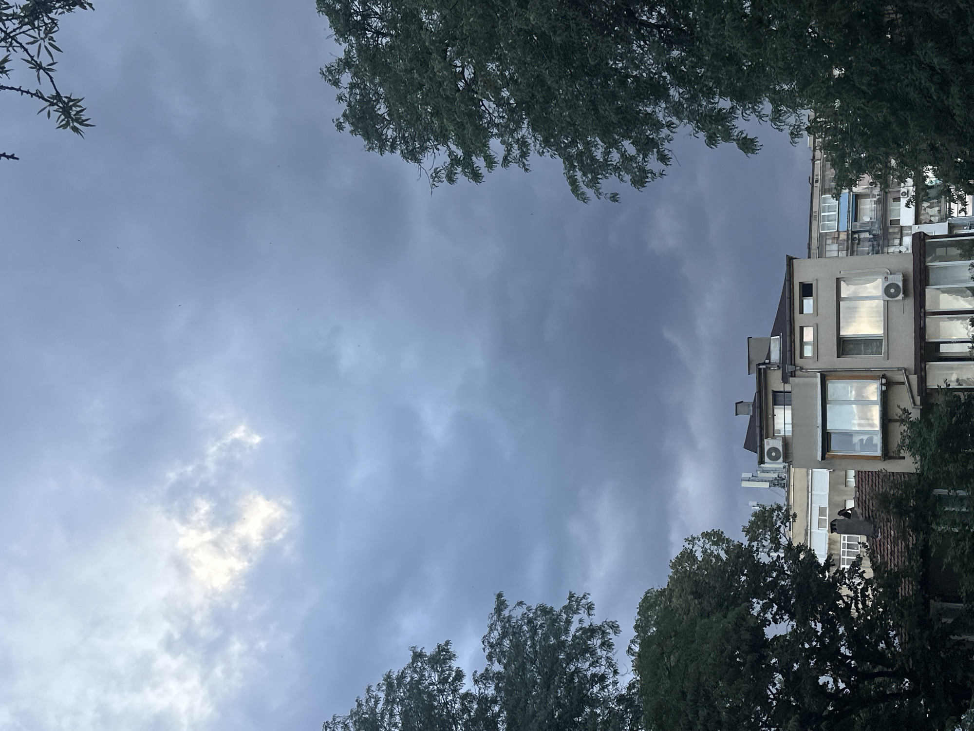 The image depicts a view of a cloudy sky above a cluster of apartment buildings, framed by trees on either side. The sky is predominantly a dark, overcast grey, with a patch of brighter light peeking through the clouds towards the upper left. Several birds are visible as small dark specks scattered across the sky.
In the foreground, parts of apartment buildings are visible. The building closest to the viewer has a brownish-red tiled roof and a lighter-colored facade with several windows. One window reflects the light from the sky. An air conditioning unit is mounted on the exterior wall below this window. Another, taller apartment building is situated behind it, with a lighter beige facade and multiple windows. This building features several antennae or communication towers on its roof. Further buildings are visible behind these, showing various architectural styles and colors, including a blue awning or section.
The trees framing the scene are lush and green, with dense foliage. The tree on the left is closer to the viewer and partially obscures the sky. The tree on the right is also prominent, with its branches and leaves reaching towards the center of the frame. A few bare branches with small leaves are visible at the very top of the image, suggesting they belong to another tree or shrub. The overall impression is a cityscape under a brooding sky, with nature intruding on the urban environment.