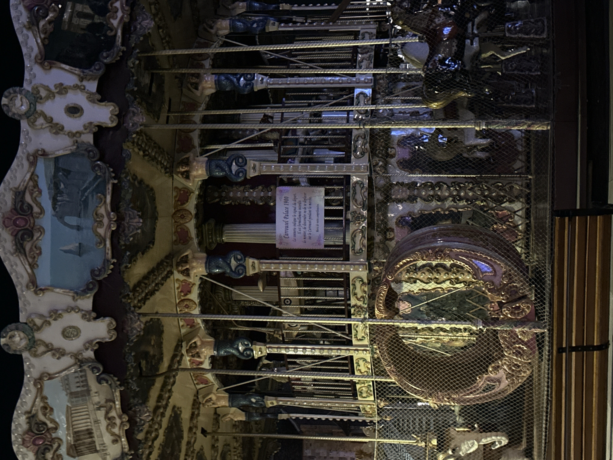 The image shows a carousel at night in the city of Honfleur, France. The carousel is elaborately decorated with ornate carvings, painted panels depicting scenes, and figures of mythological creatures and animals. The carousel is illuminated, casting a warm glow on the surrounding area. In the foreground, a wooden bench is visible, suggesting a public space. The carousel itself features several horses and other rideable figures. A sign on the carousel reads "Carrousel Palace 1900," indicating its historical significance and name. The overall impression is one of a charming and nostalgic attraction, likely a popular spot for both locals and tourists.